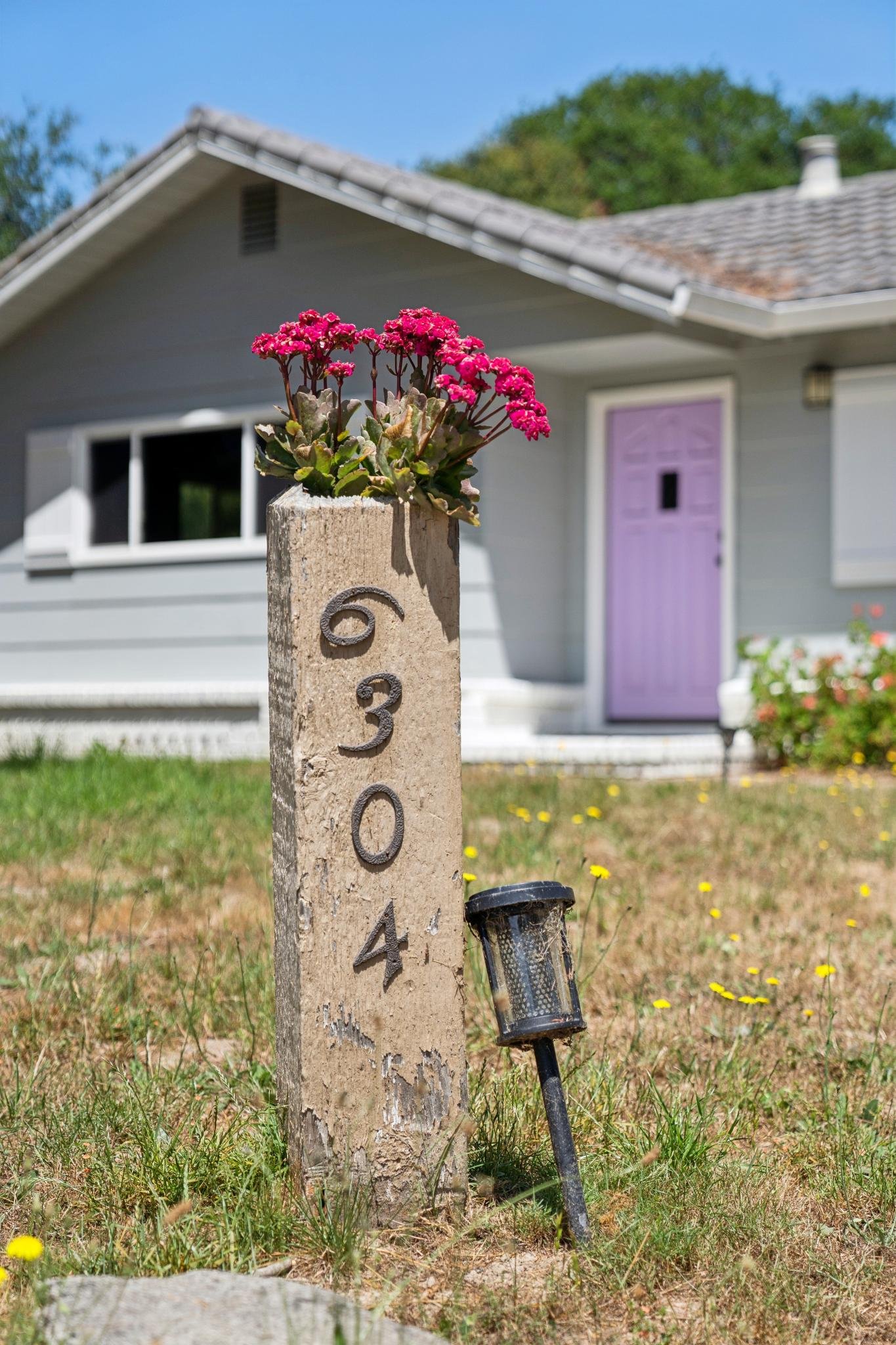 A wooden post displaying house numbers '60340' with pink flowering plant on top, a lantern light missing, in front of a gray house with purple door, surrounded by grass and small yellow flowers.
