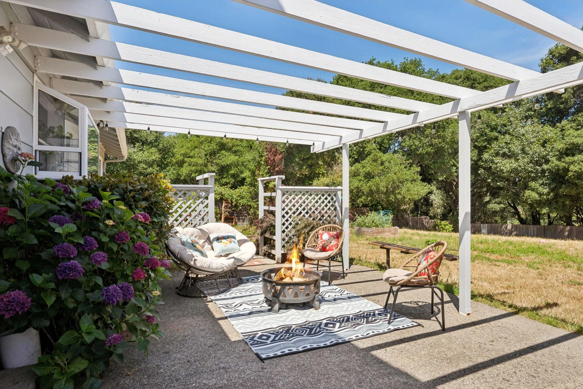 An outdoor patio area with a white pergola, garden furniture including chairs and a fire pit, surrounded by lush green trees and flowering bushes, with a blanket on the ground and a string of lights hanging from the pergola.