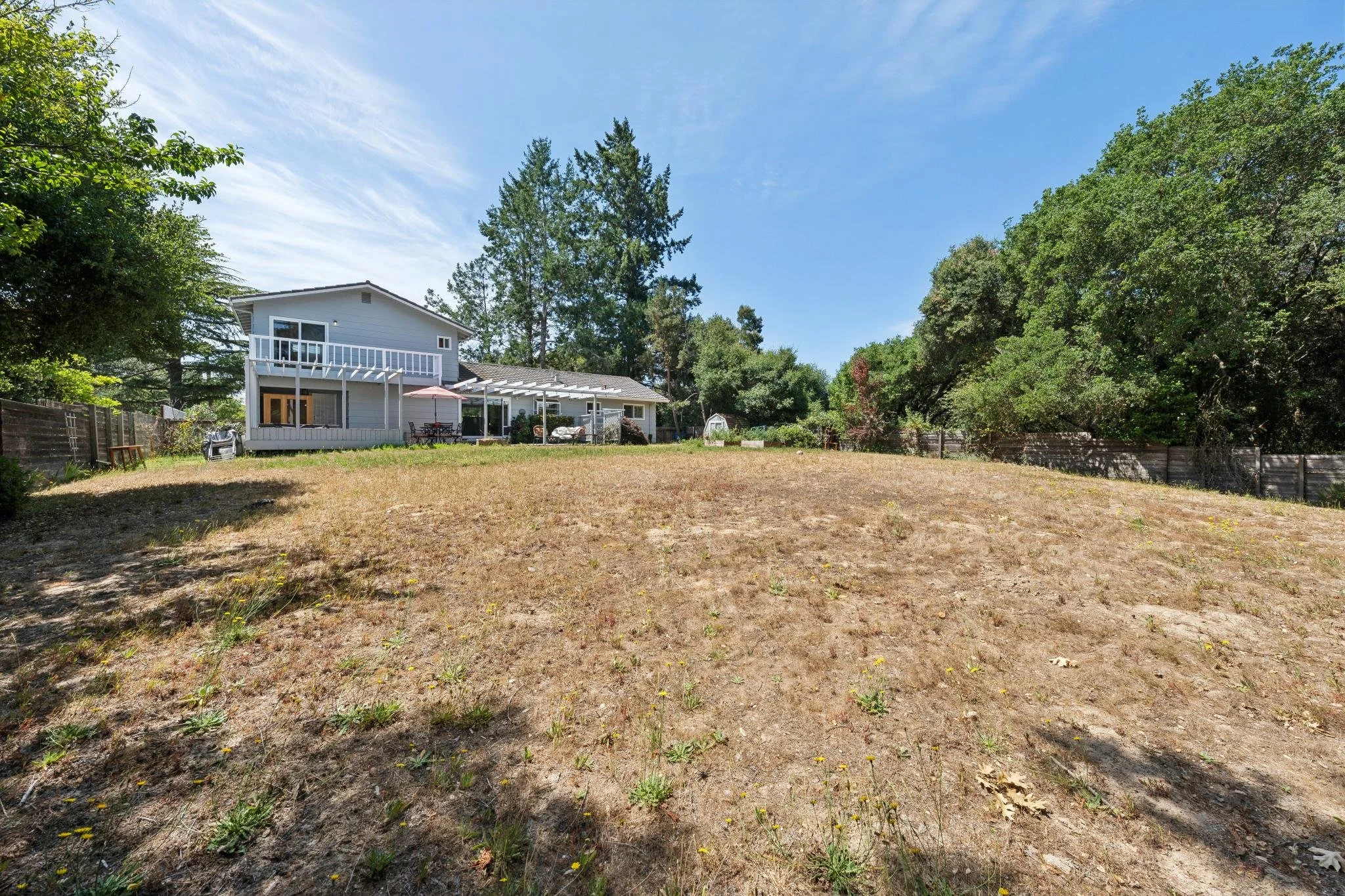 A backyard yard with a large open grassy area, a two-story house in the background with an upper balcony, surrounded by tall trees and a clear blue sky.