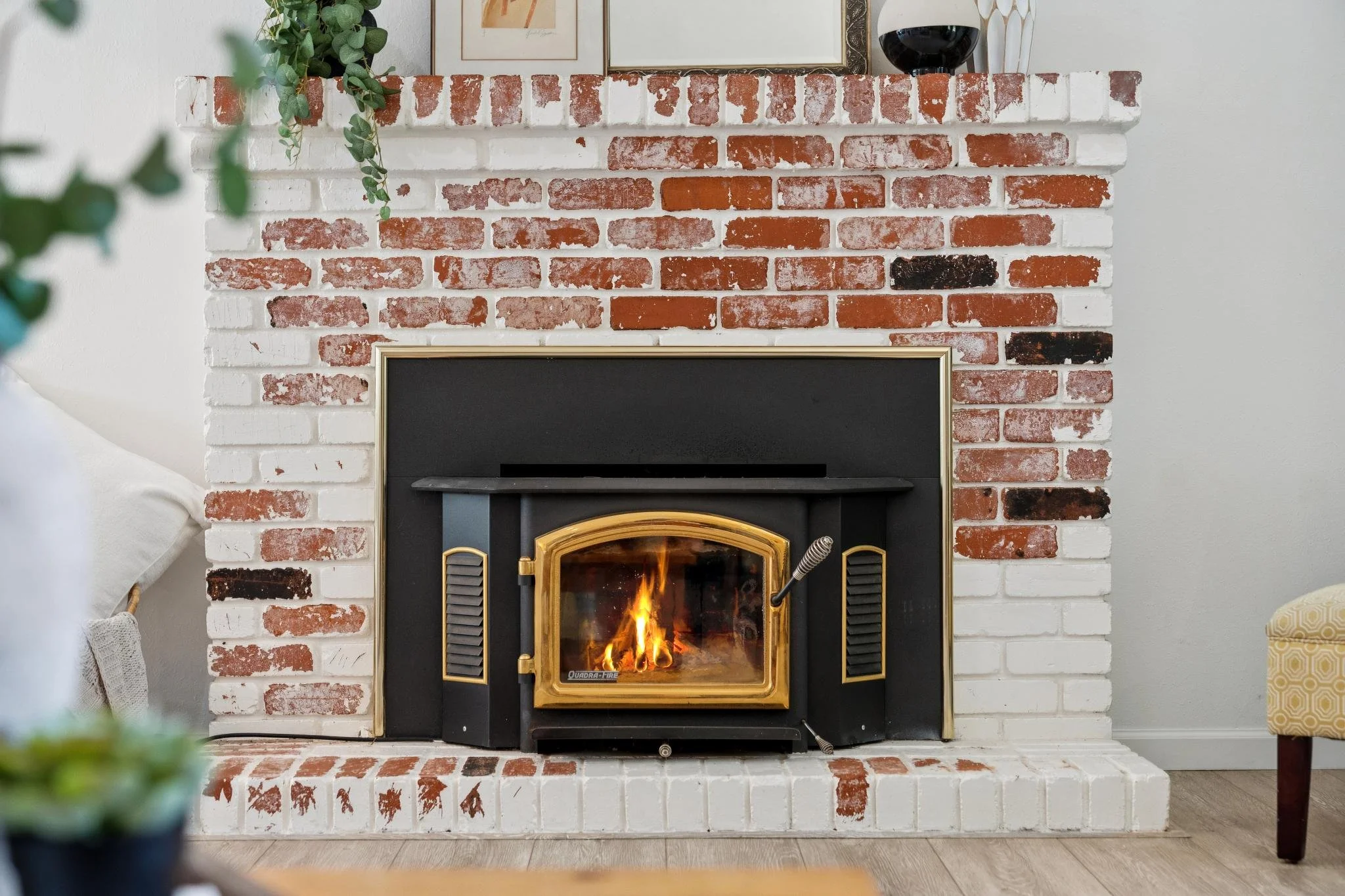 A fireplace with a burning fire, set within a white and red brick surround. Decor items are placed on the mantle, including a framed picture, a plant, and a glass of wine.