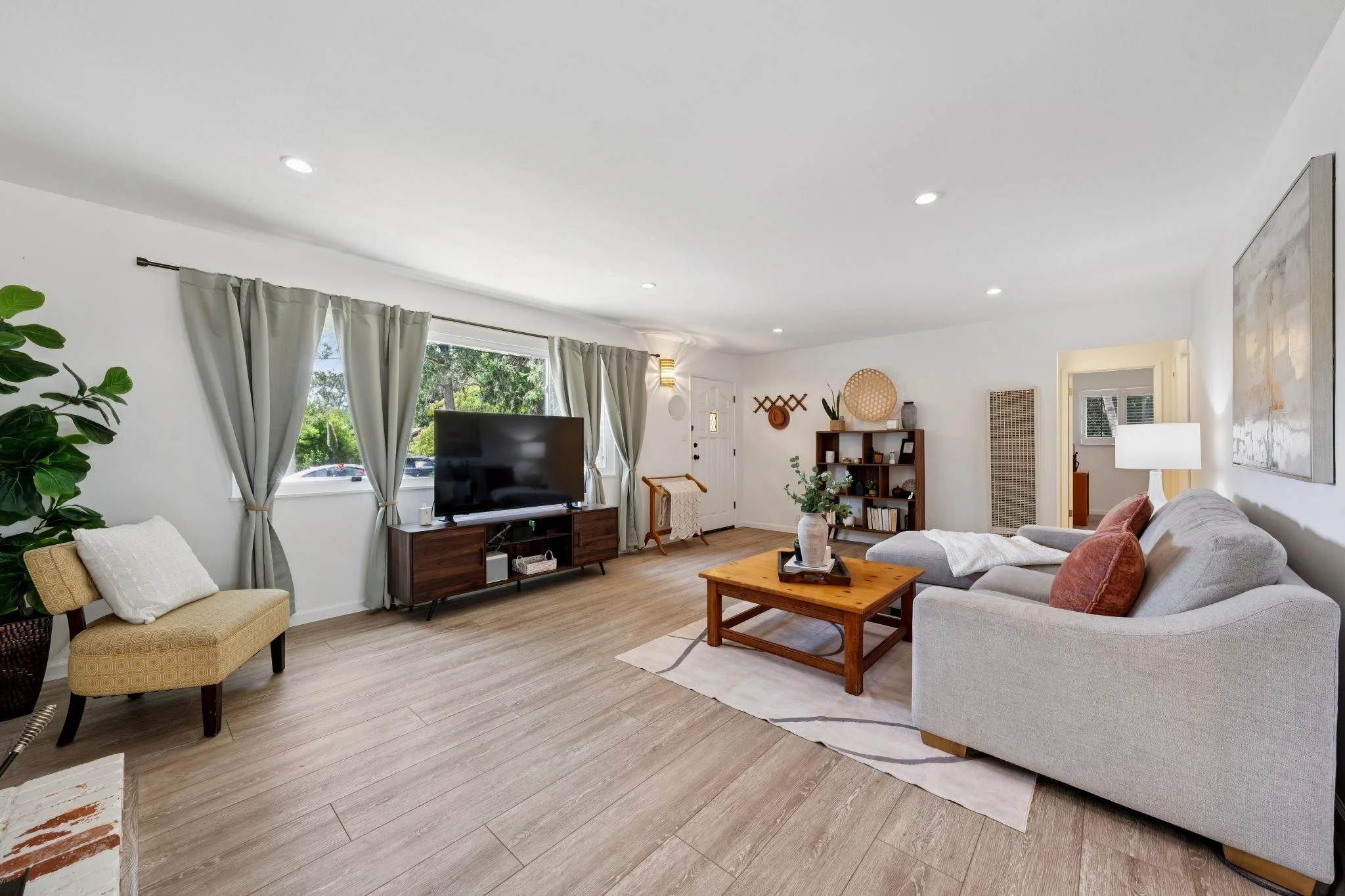 Living room with white walls, light hardwood floor, a gray sofa with pink pillows, a wooden coffee table, a large window with curtains, a TV on a wooden stand, a shelving unit with decor, and a large abstract painting.