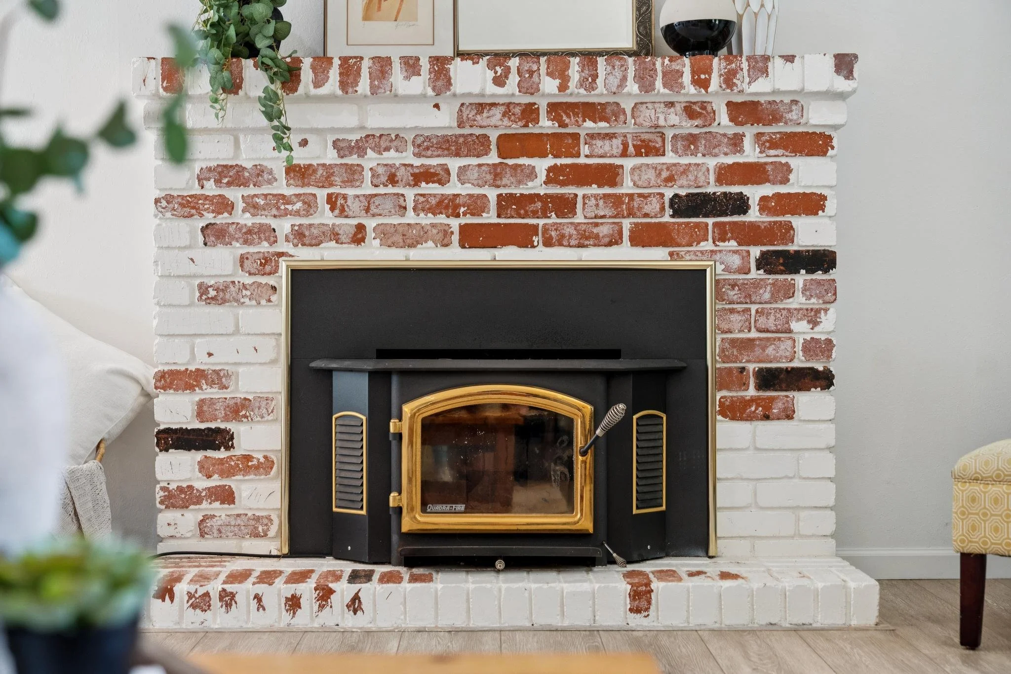 A brick fireplace painted white around a black and gold wood stove, decorated with framed artwork, a green plant, and a glass of red wine on top.