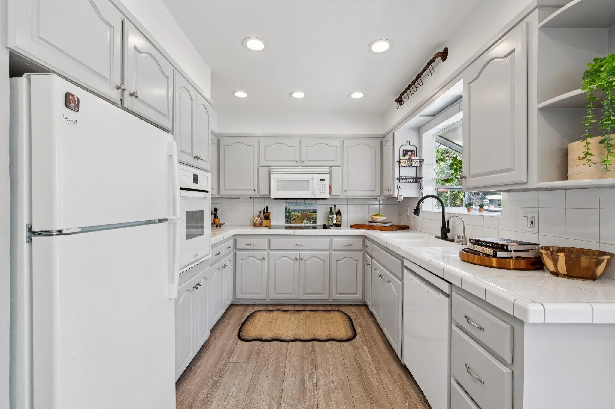 A bright white kitchen with white cabinets, a white countertop, and a window. There are various small kitchen items, bottles, and plants on the counter and shelves. The floor is wood, and a small beige rug is in front of the sink.