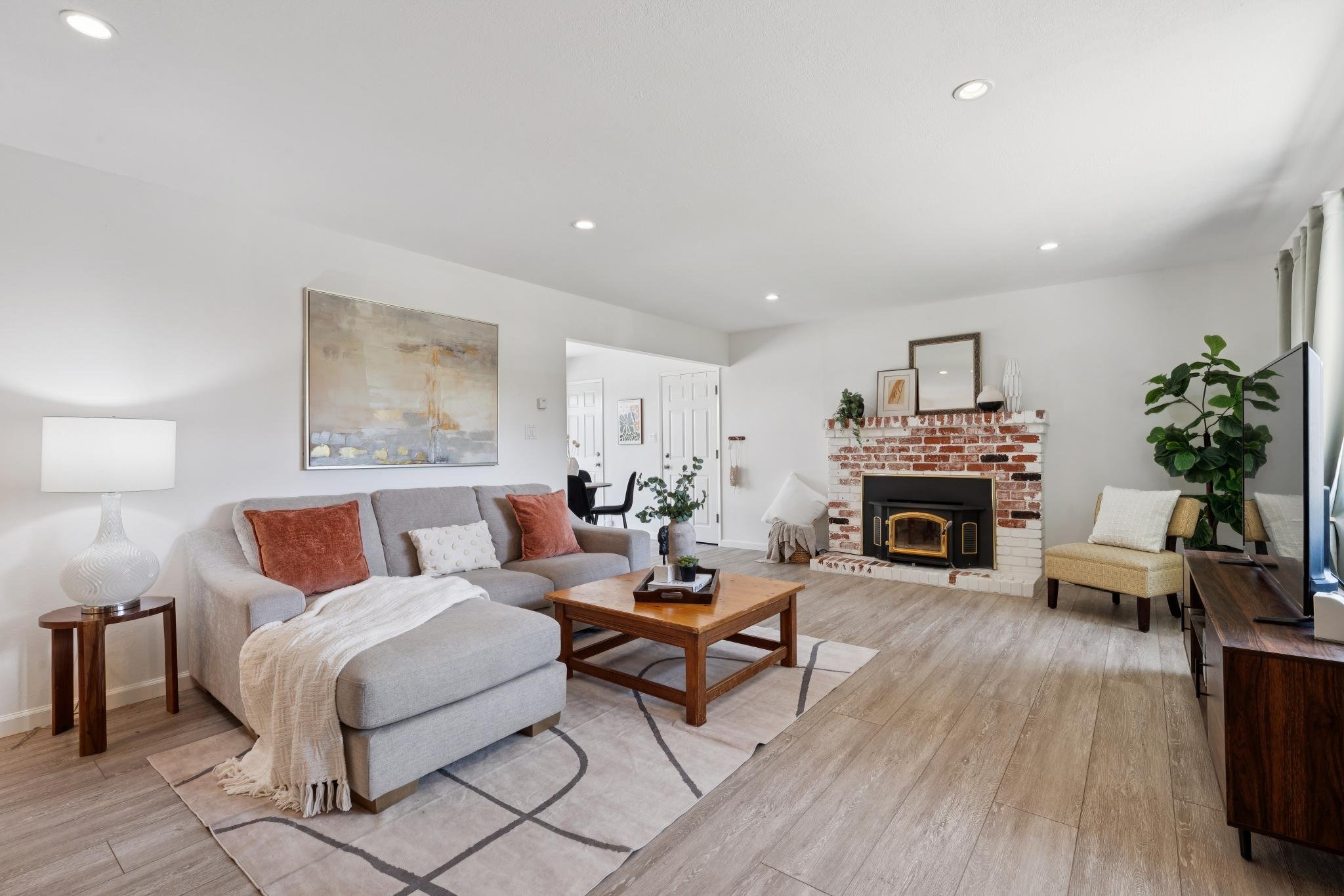 Living room with beige sofa, orange and white pillows, a wooden coffee table, fireplace with brick surround, wall art, potted plants, and a TV.