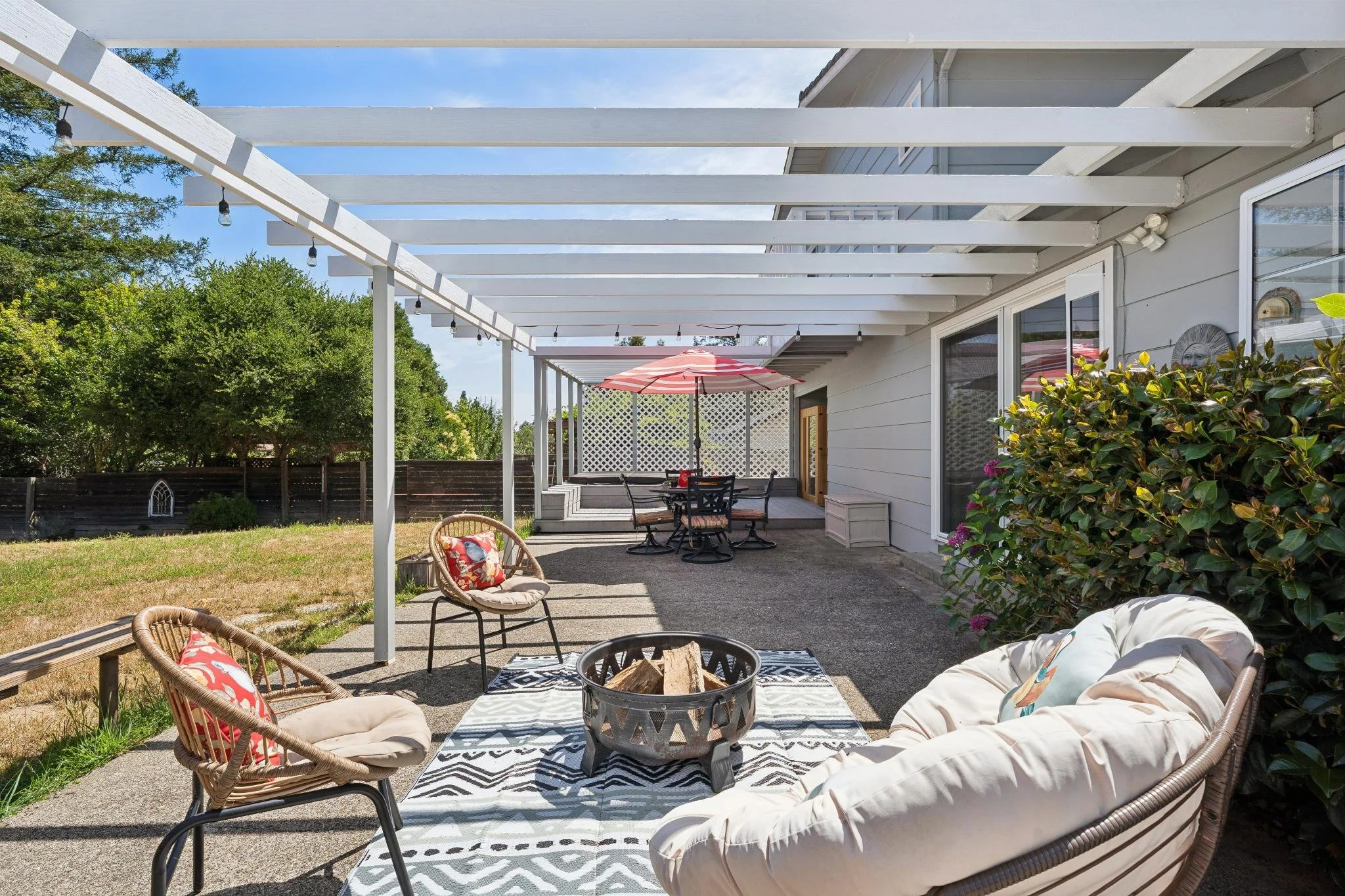 A backyard patio with outdoor furniture under a white pergola, including a fire pit, chairs with patterned pillows, and a table with an umbrella, surrounded by greenery and a wooden fence.