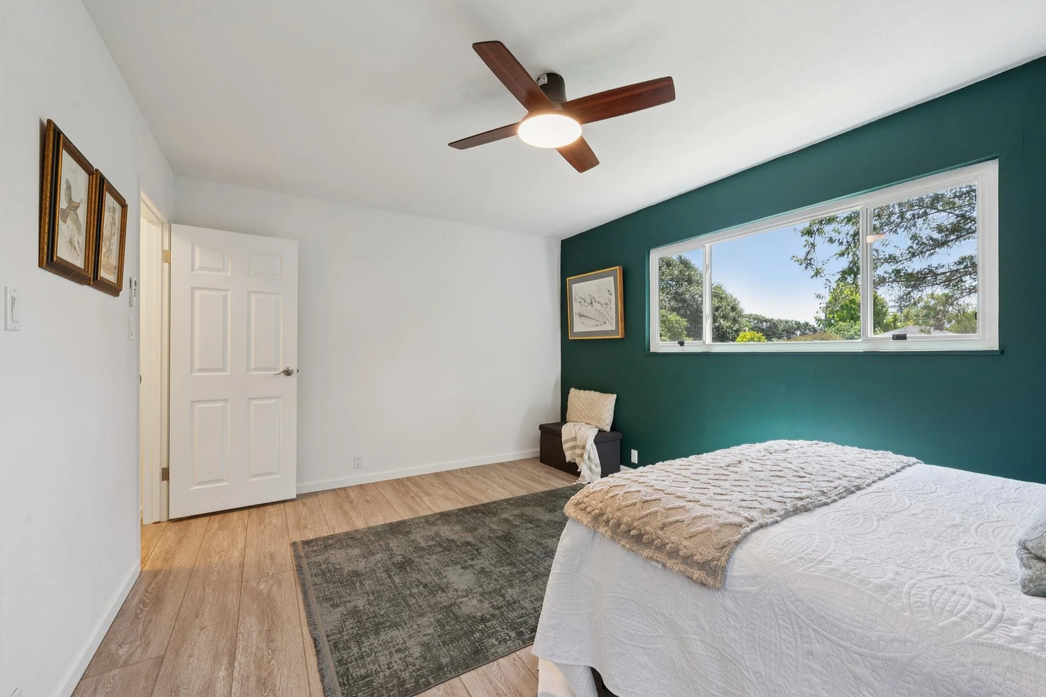 Interior of a bedroom with a white wall and a dark green accent wall. A large window shows trees outside. There is a bed with white bedding and a beige blanket, a small dark wood nightstand with a cream-colored pillow and a beige and white blanket. T