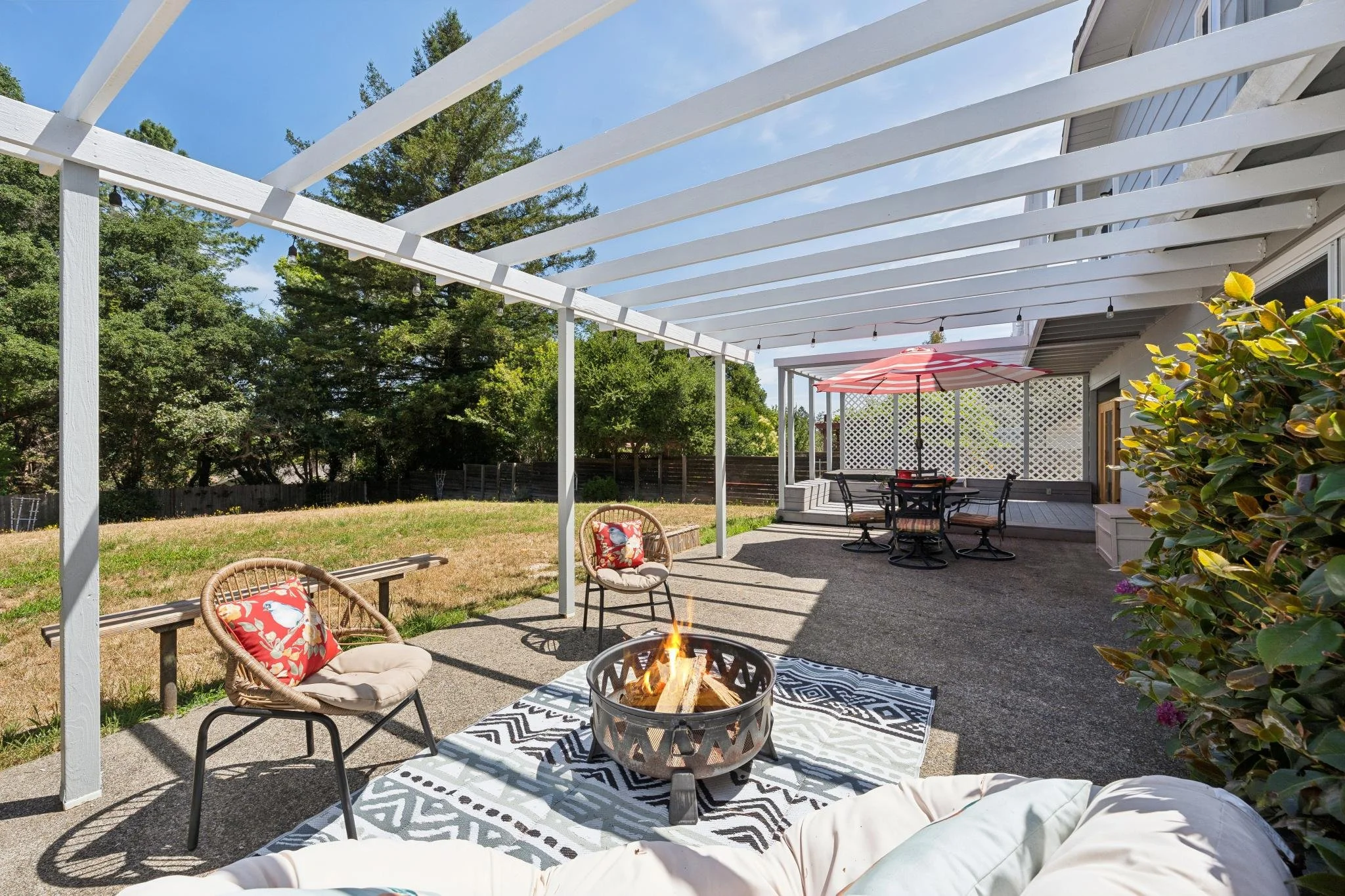 Backyard patio with white pergola, outdoor furniture including chairs with cushions, a round fire pit, a table with an umbrella, and green trees in a sunny setting.