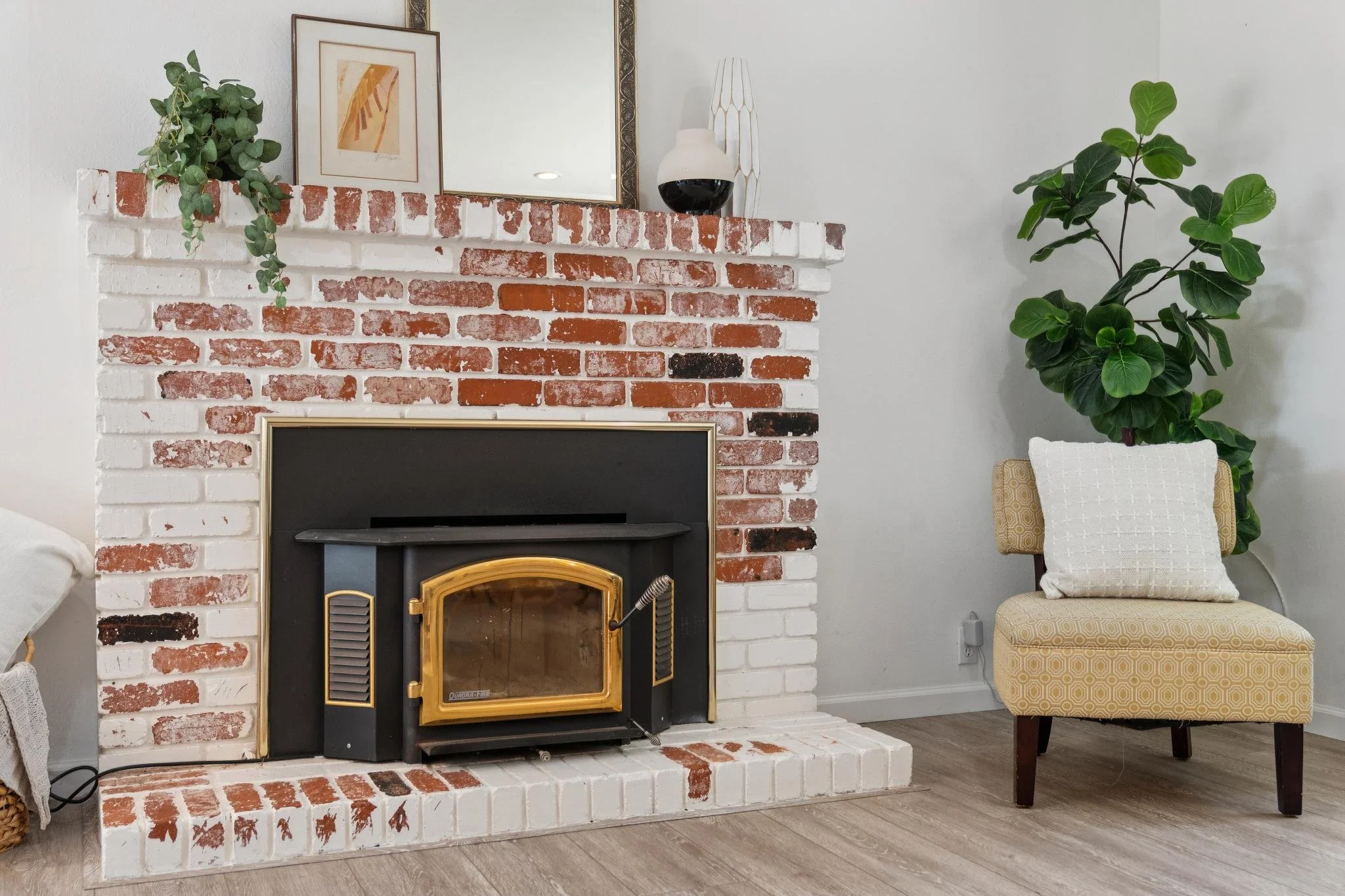A fireplace with red and white bricks, a framed artwork, a mirror, a black and white vase, and a large green houseplant in a living room.