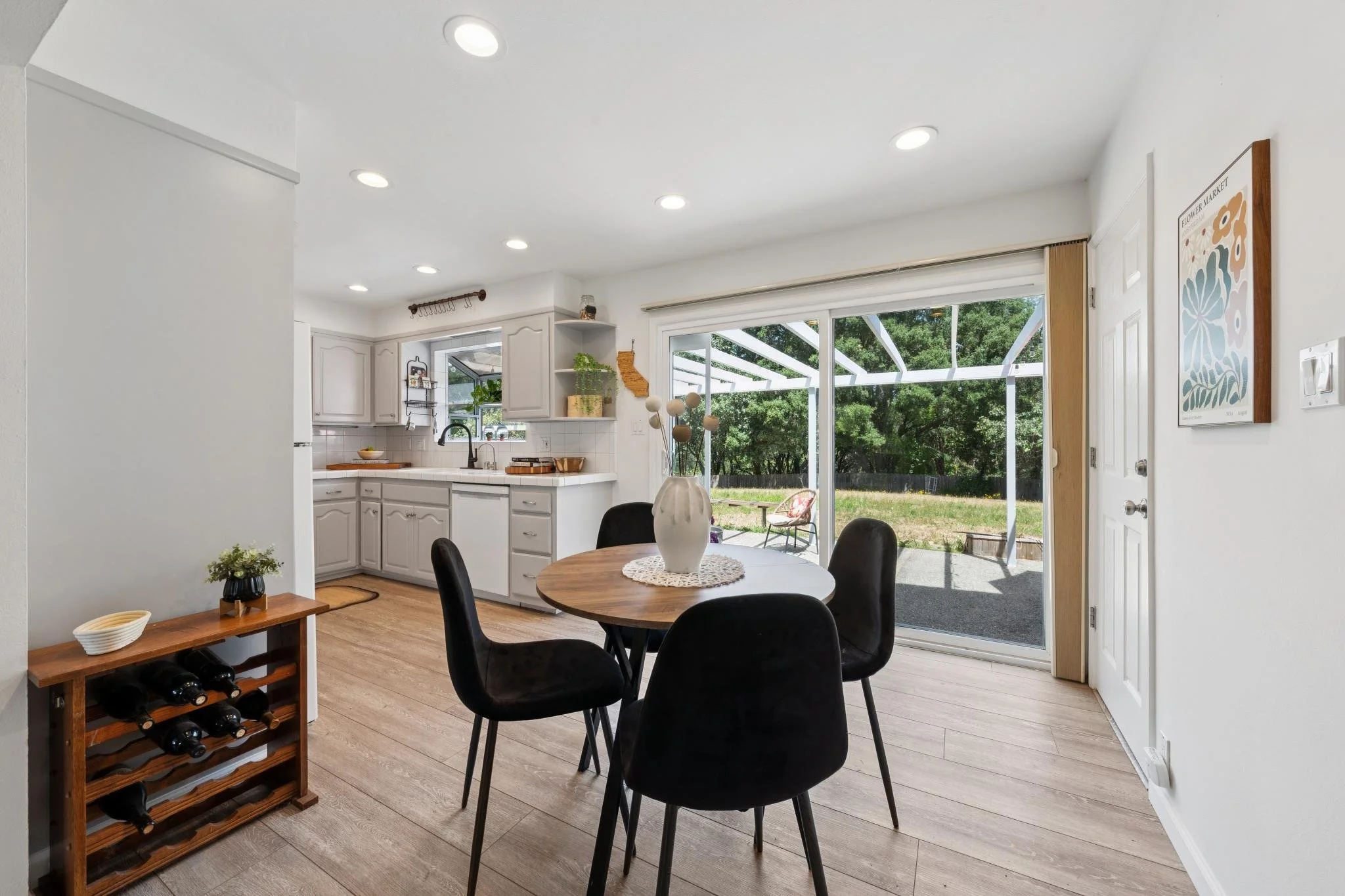 Interior view of a bright dining kitchen with sliding glass door leading outside, round dining table with black chairs, white cabinets, wooden floor, and decorative items.