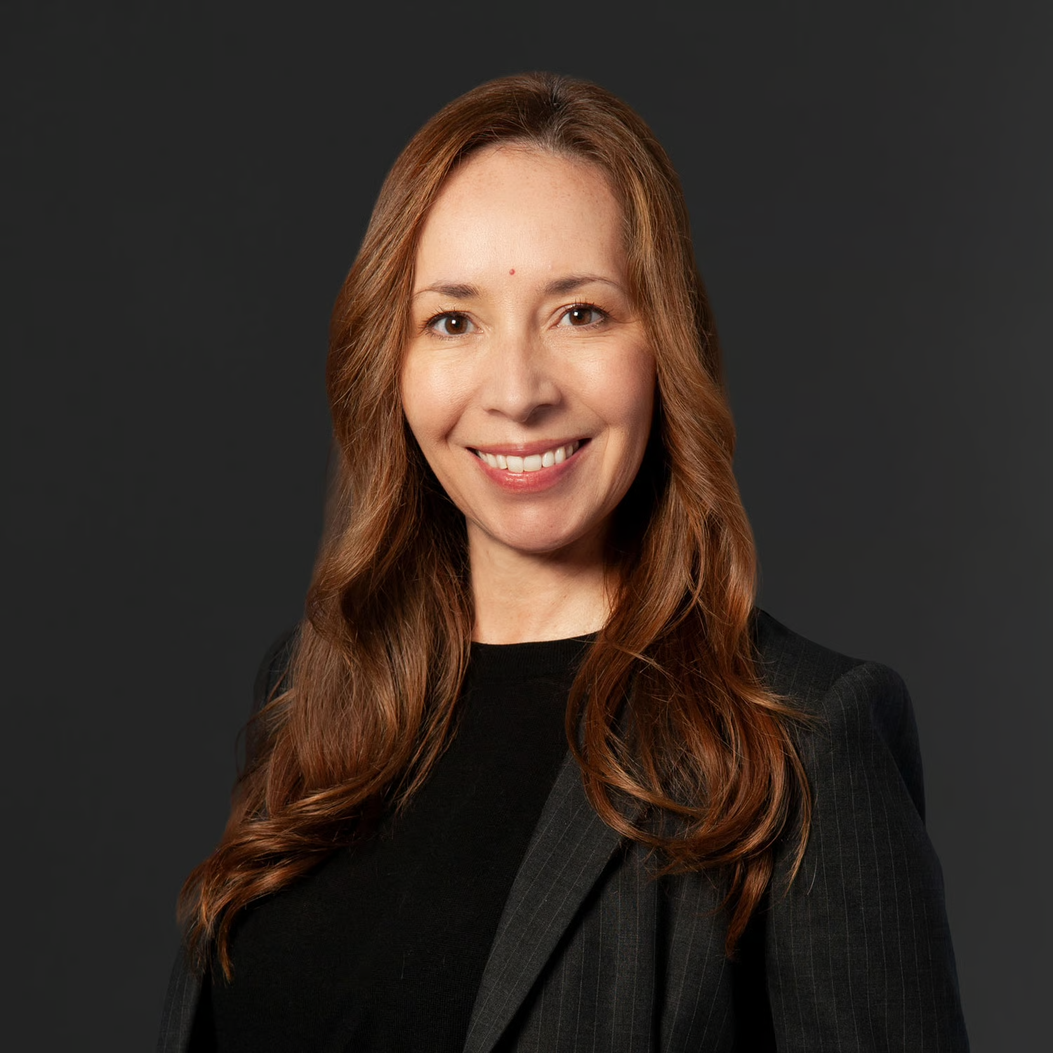 A professional woman with long wavy red hair, wearing a dark professional outfit, smiling against a plain dark background.