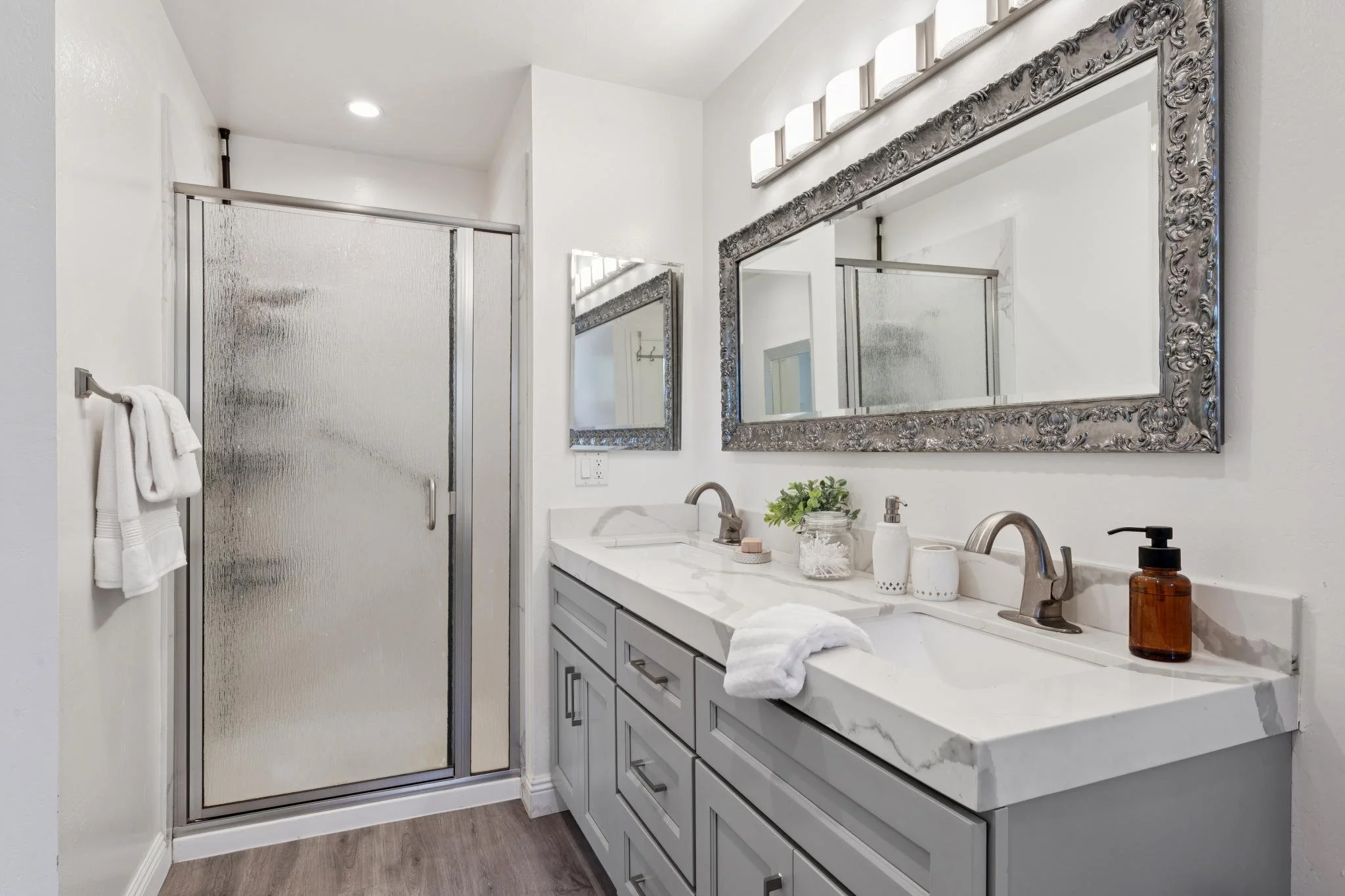 Modern bathroom with white vanity, silver-framed mirrors, frosted glass shower, and decorative accessories.