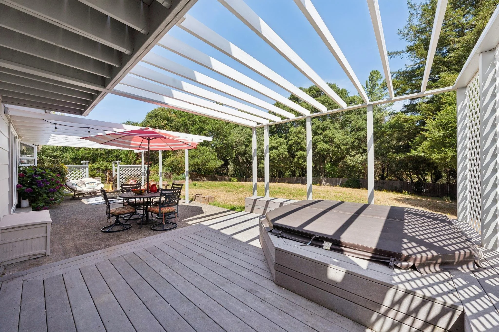 Backyard patio with grey wooden decking, a covered pergola, patio furniture including a round table with chairs, an umbrella, and a hot tub, surrounded by green trees and a lawn.