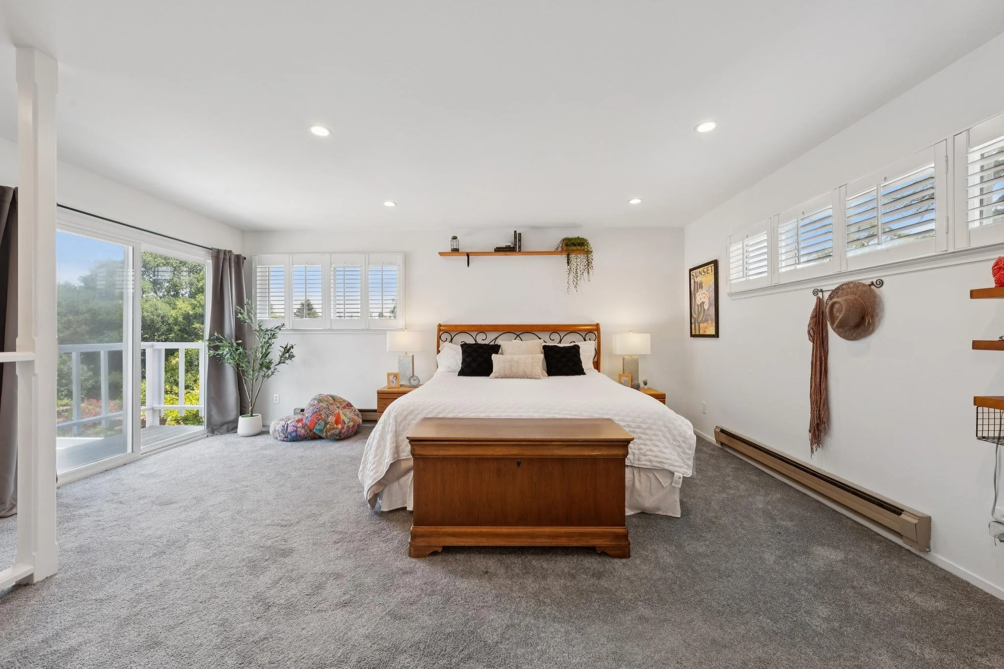 Bedroom with a wooden bed frame, white bedspread, black and beige pillows, bedside lamps, a wall shelf with decorations, a framed sunset poster, and large windows with white shutters and gray curtains. There is a plant near the window and two colorfu