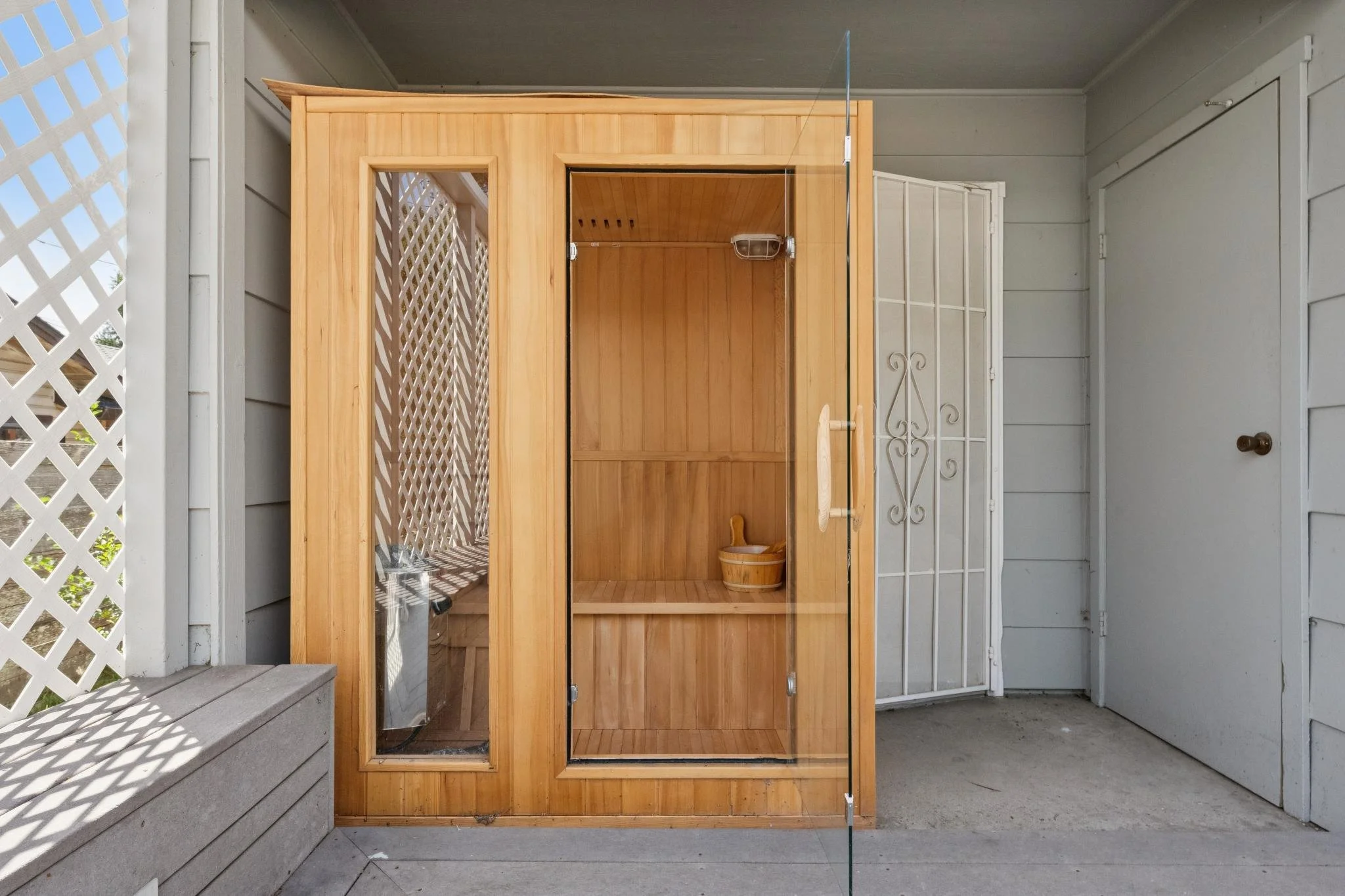 A wooden outdoor sauna with a glass door, situated on a porch with gray wooden siding, a white lattice panel, and a white metal gate.