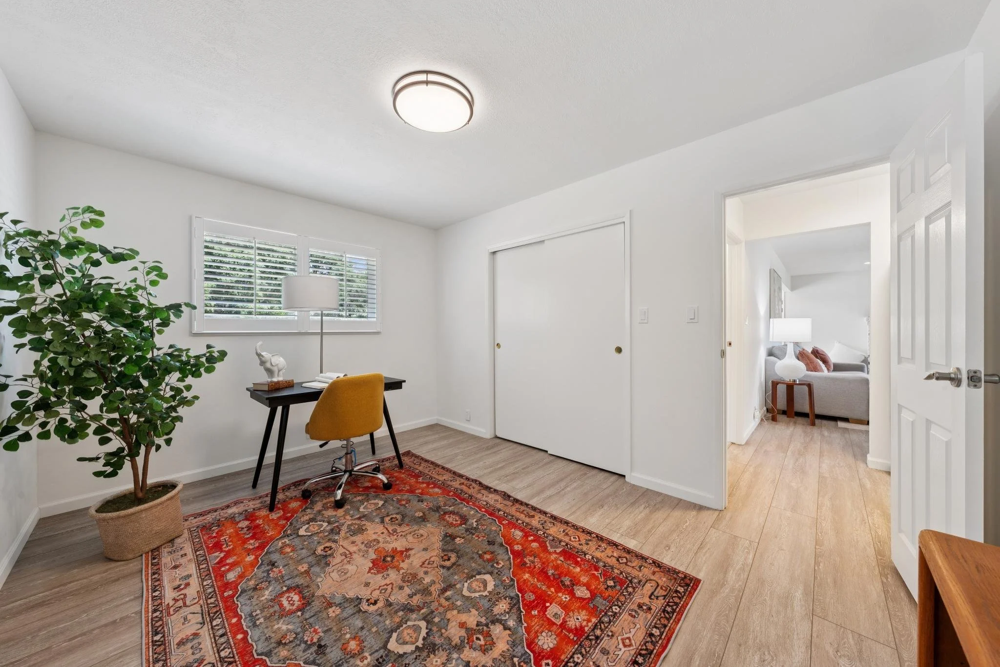 An interior view of a room with white walls, a window with shutters, a potted plant, a desk with a yellow chair, a white table lamp, and a decorative area rug.