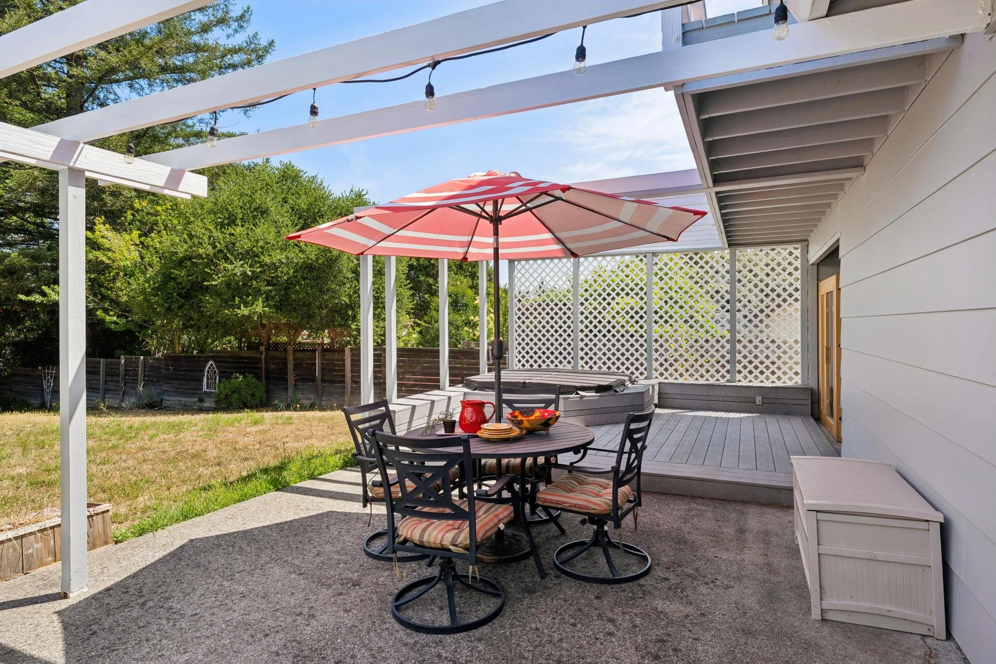 A backyard patio with a round table and six chairs, a red and white striped umbrella, and string lights hanging overhead. There's a grassy yard and trees in the background, with a fence surrounding the yard.