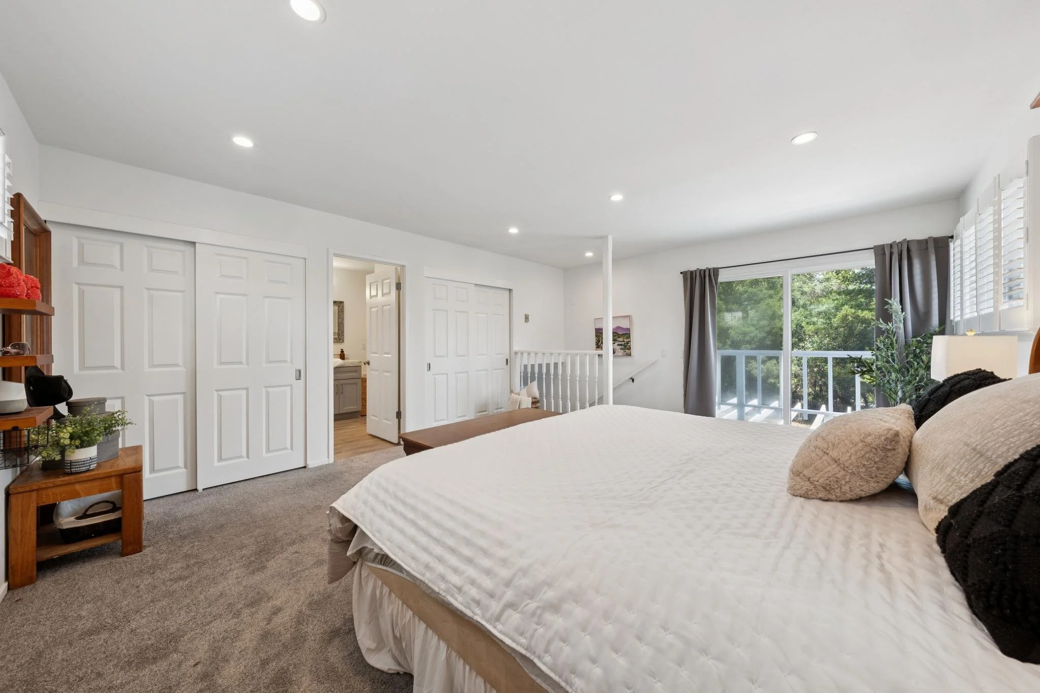 A bedroom with a bed, beige bedding, pillows, a window with gray curtains, and a sliding door closet. There's a small wooden nightstand with plants and decorative items.