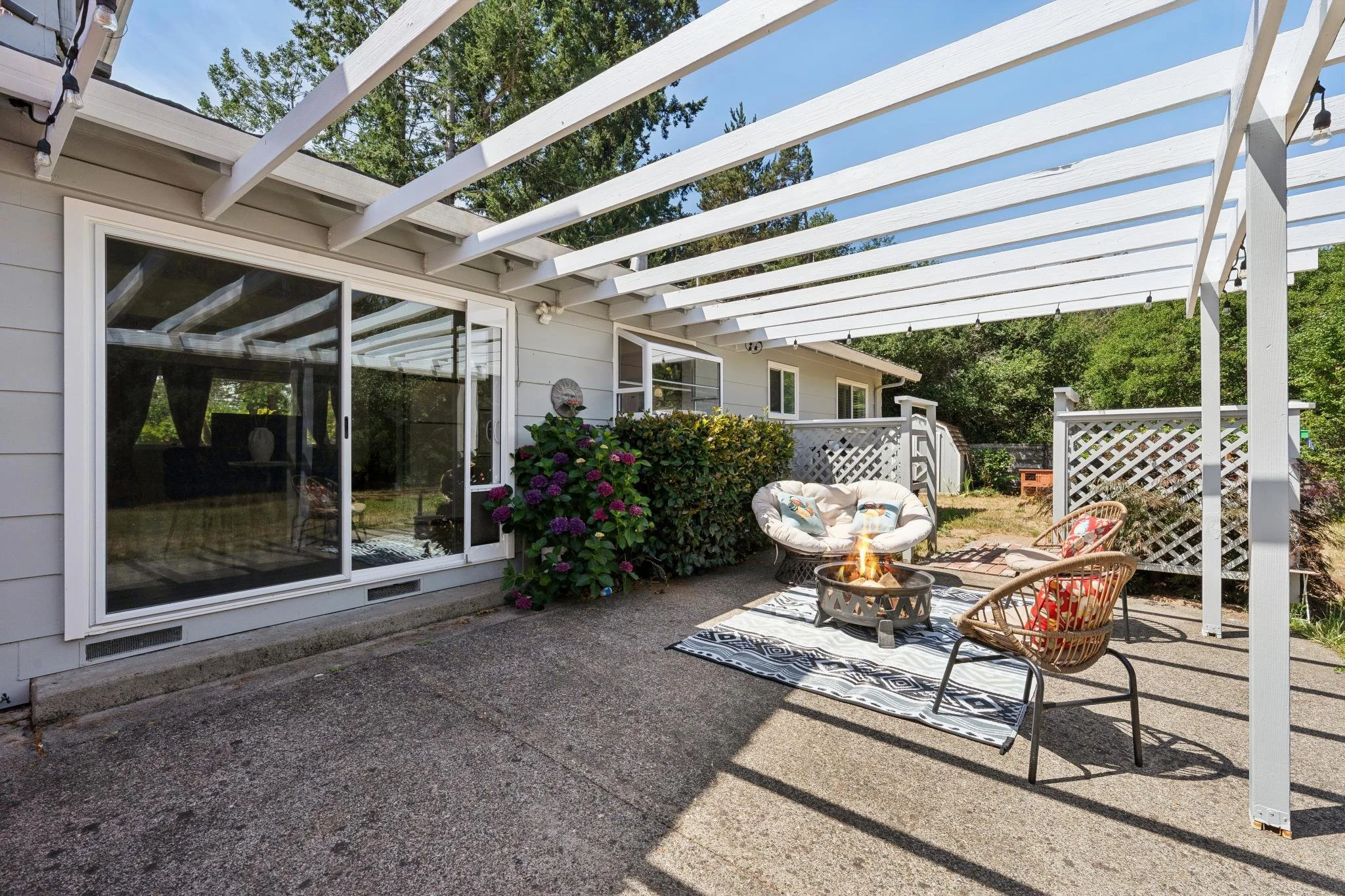 Backyard patio area with white painted pergola, sliding glass door, outdoor furniture, potted plants, and a fire pit on a patterned outdoor rug.