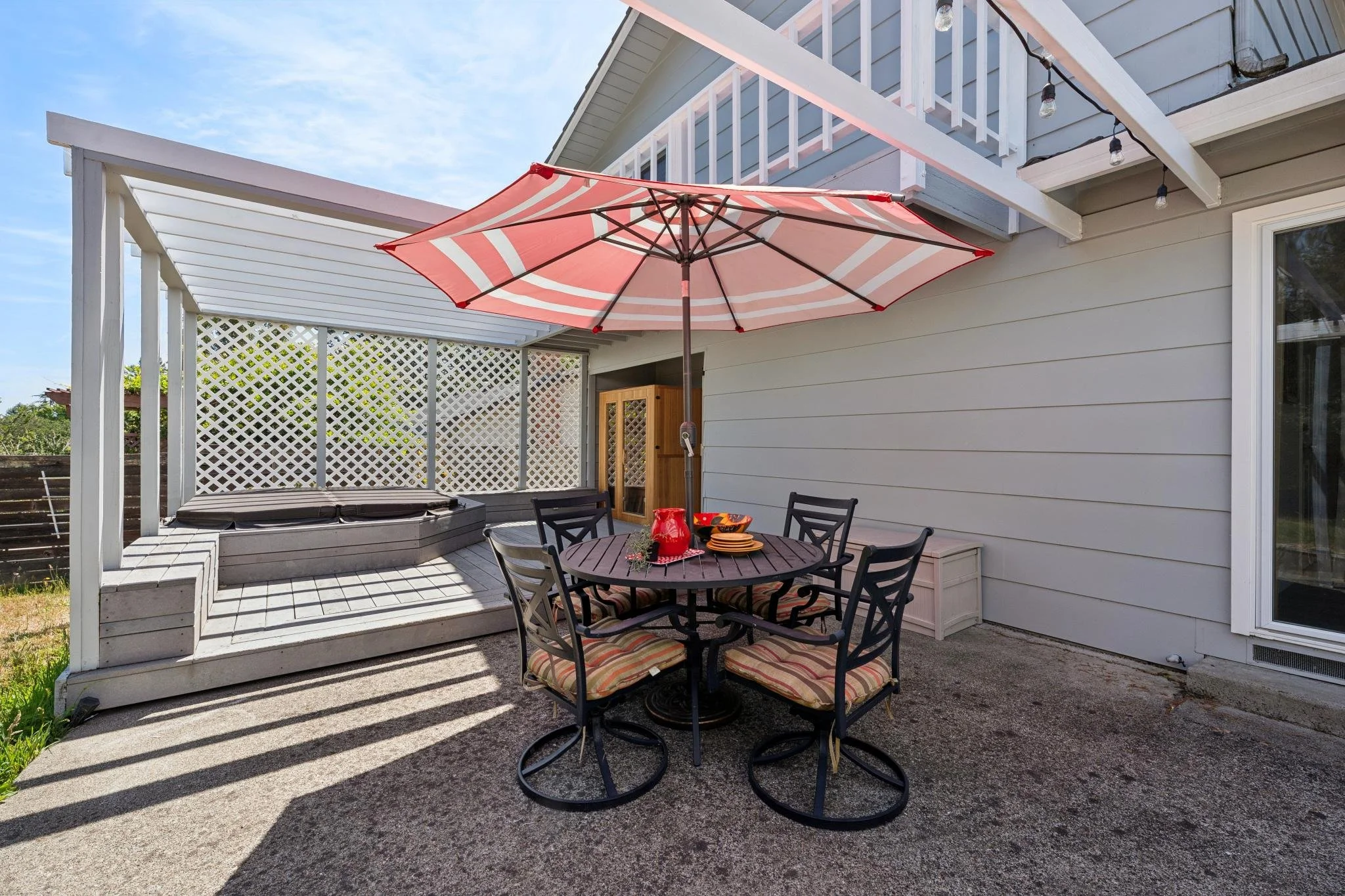 An outdoor patio area with a round table and six chairs, under a large umbrella with pink, white, and red stripes. The table is set with bowls, a red pitcher, and a cloth. The patio is attached to a house with gray siding and white trim, with string 