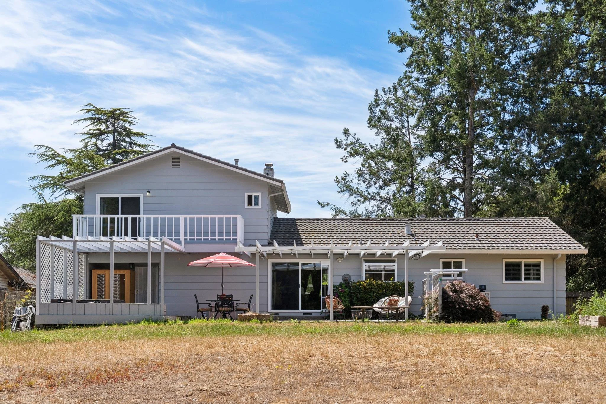 Backyard view of a two-story house with a balcony and outdoor furniture, surrounded by large trees and a partly cloudy sky.