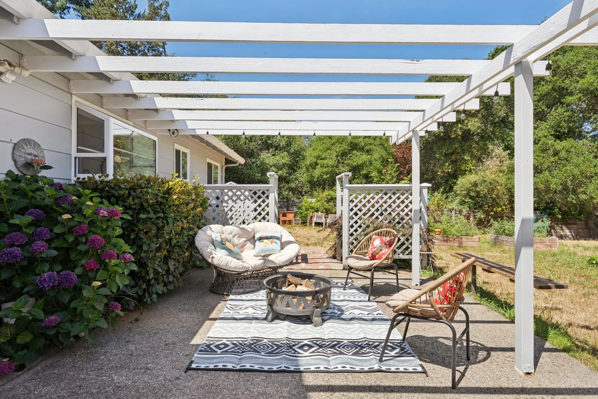 An outdoor patio area with white pergola, seating, a patterned rug, and surrounding greenery.