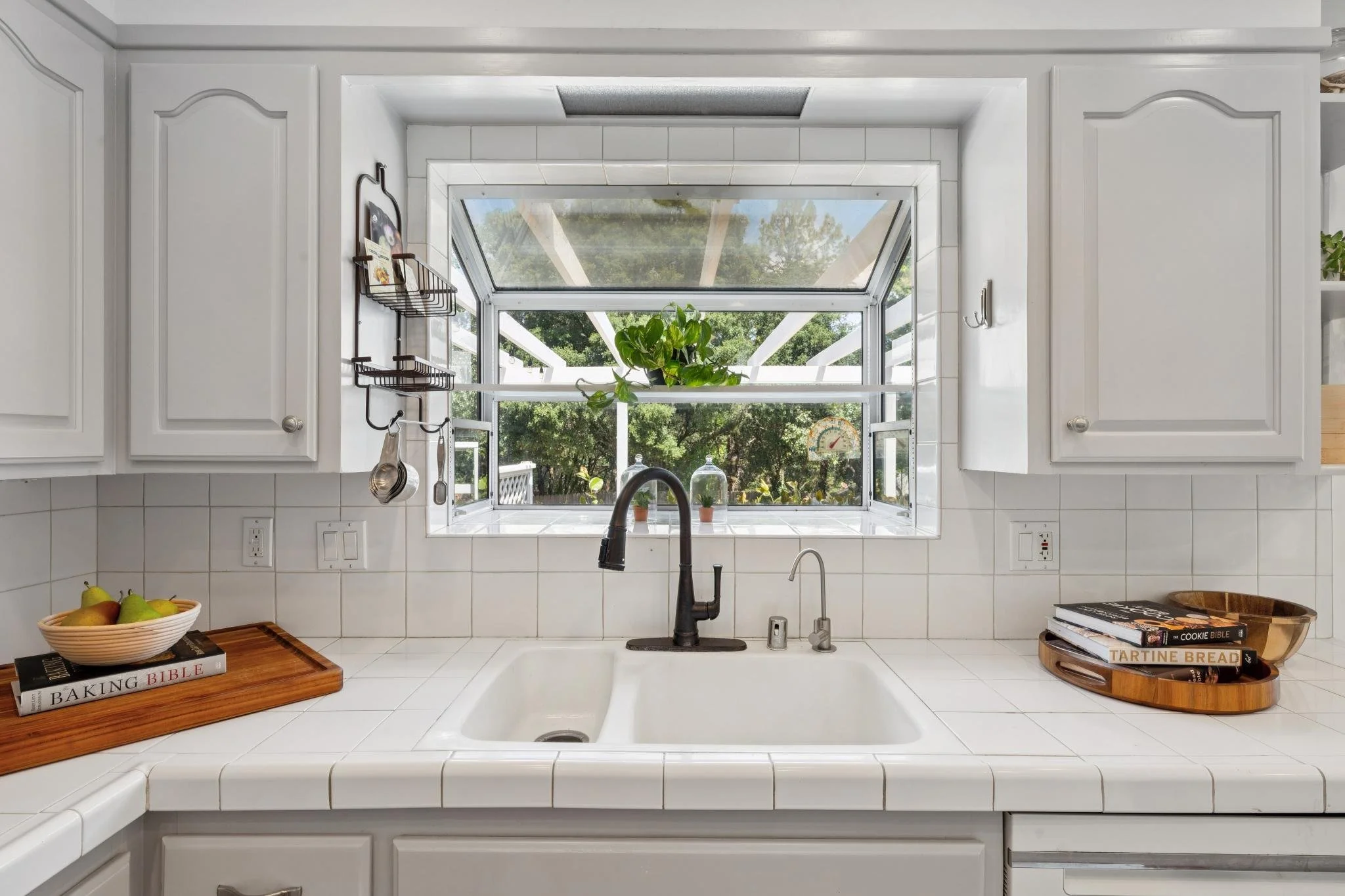 A kitchen with white cabinets, a window above the sink, and a black faucet. On the counter, there is a bowl of pears and a wooden tray with baking books, including 'Baking Bible' and 'The Cookie Bible'.