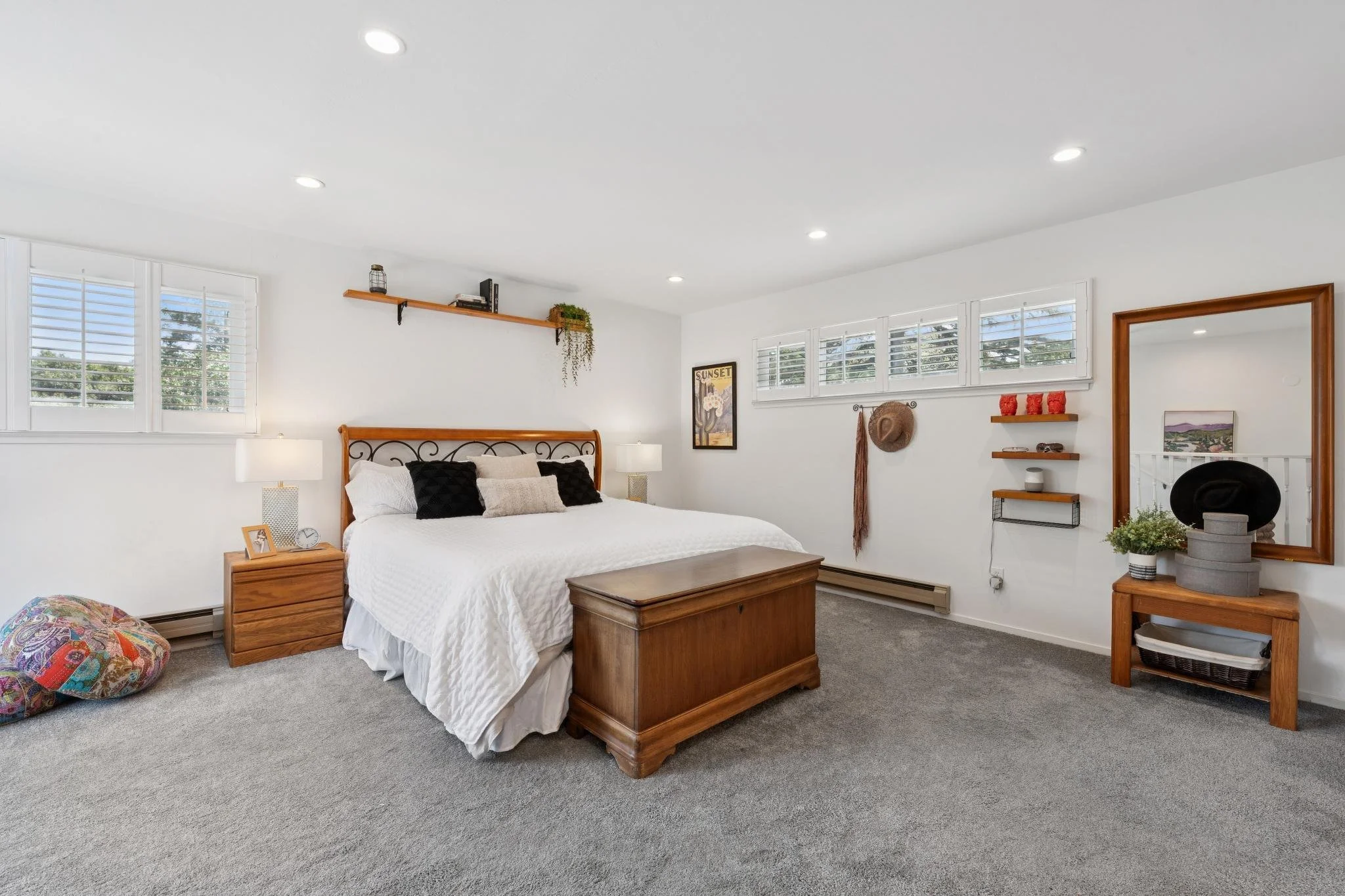 A bedroom with white walls, a bed with white bedding and black pillows, two wooden nightstands with lamps, a wooden bench at the foot of the bed, and a large mirror on the wall. There are three windows with white shutters, a small shelf with decorati