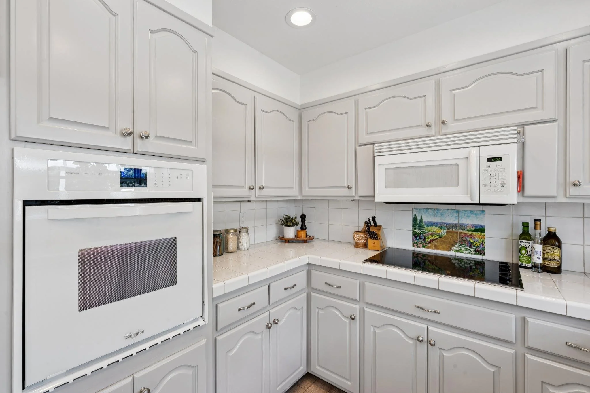 White kitchen cabinets, built-in oven, microwave, and stove, with various jars, bottles, and kitchen tools on the countertops.