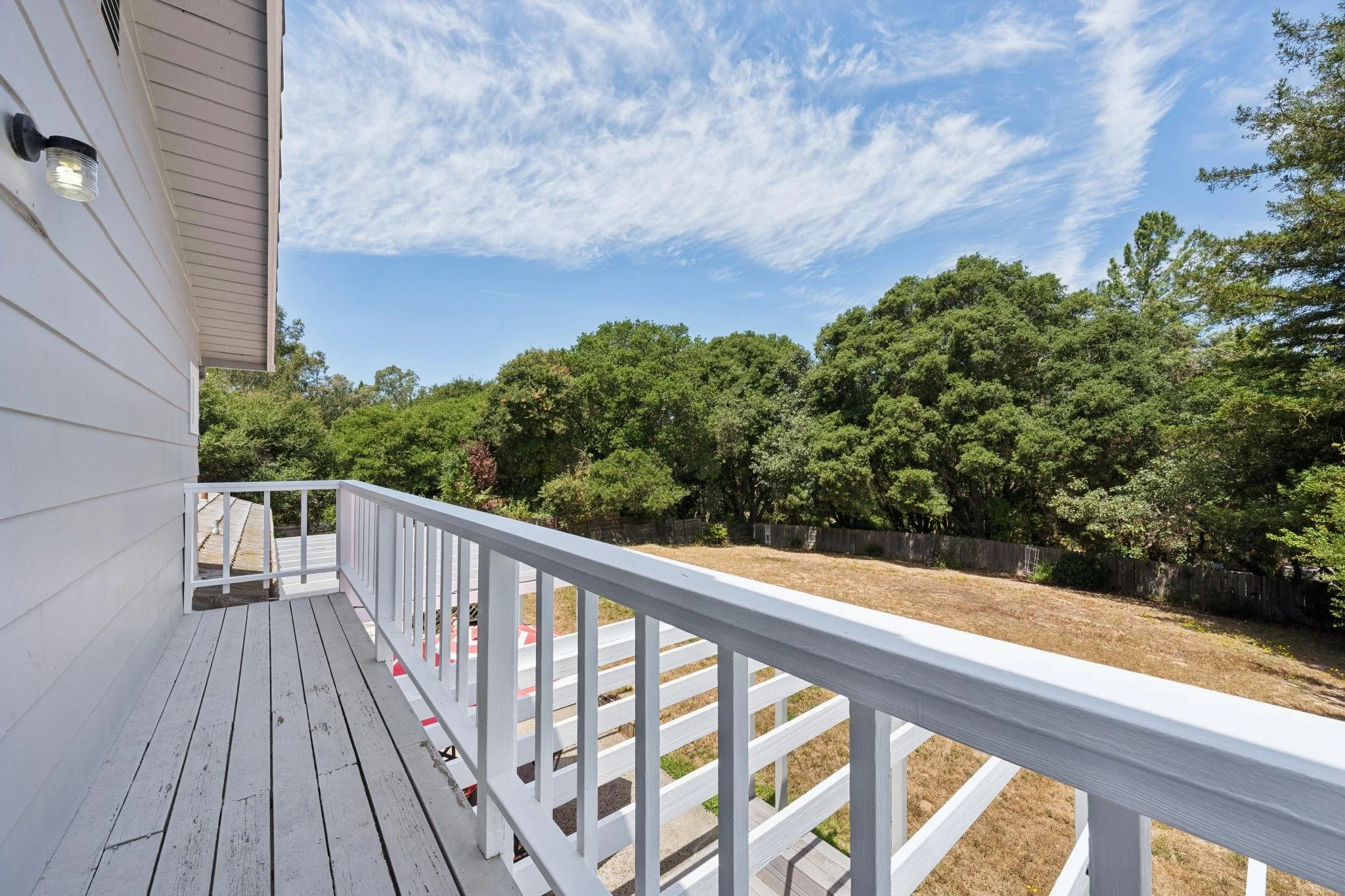 View from a white wooden balcony with railing, looking out over a yard with trees and a wooden fence, under a blue sky with wispy clouds.