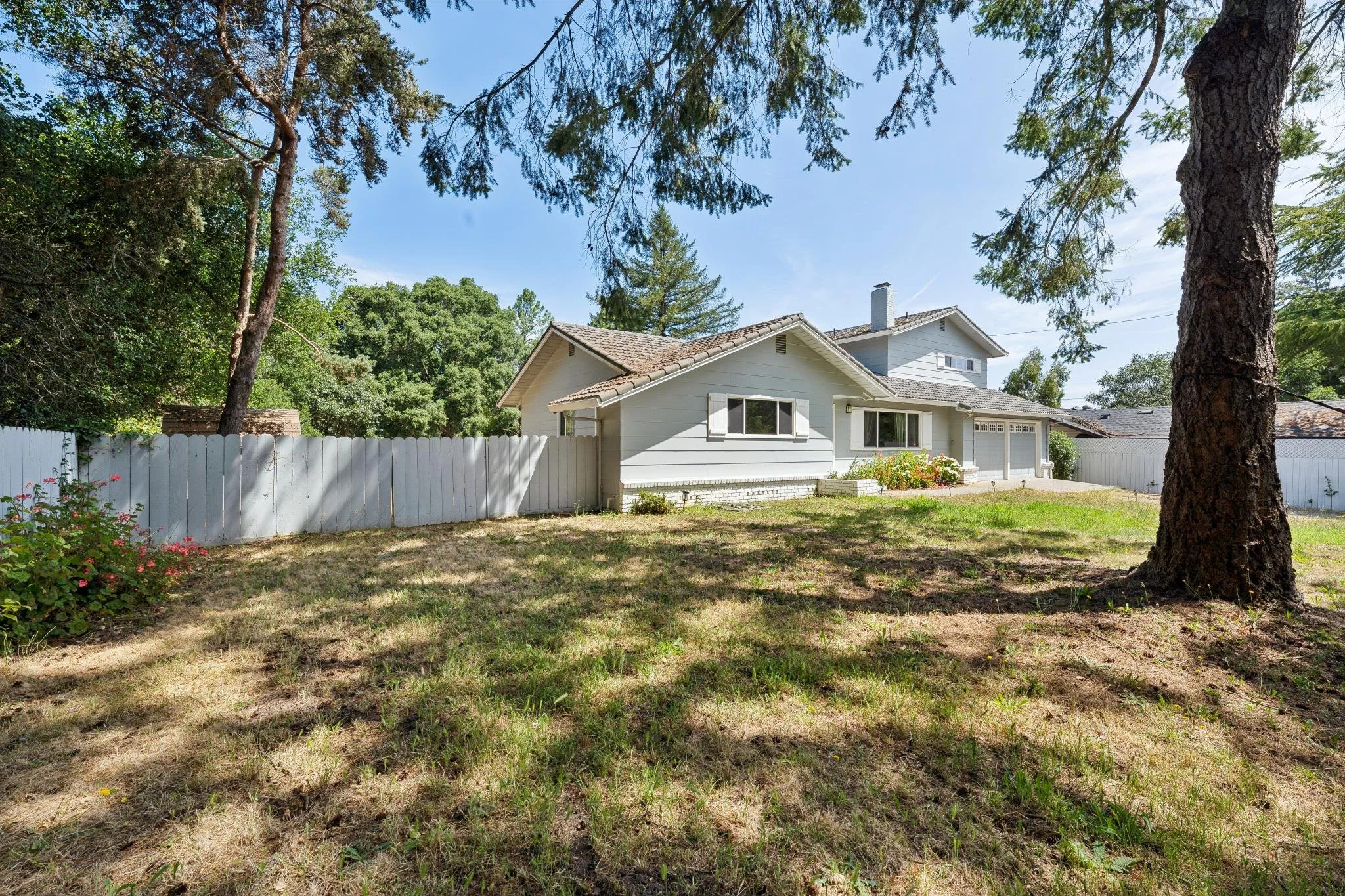 A backyard scene with a white house, tall trees, a white picket fence, and a small flower garden under a clear blue sky.