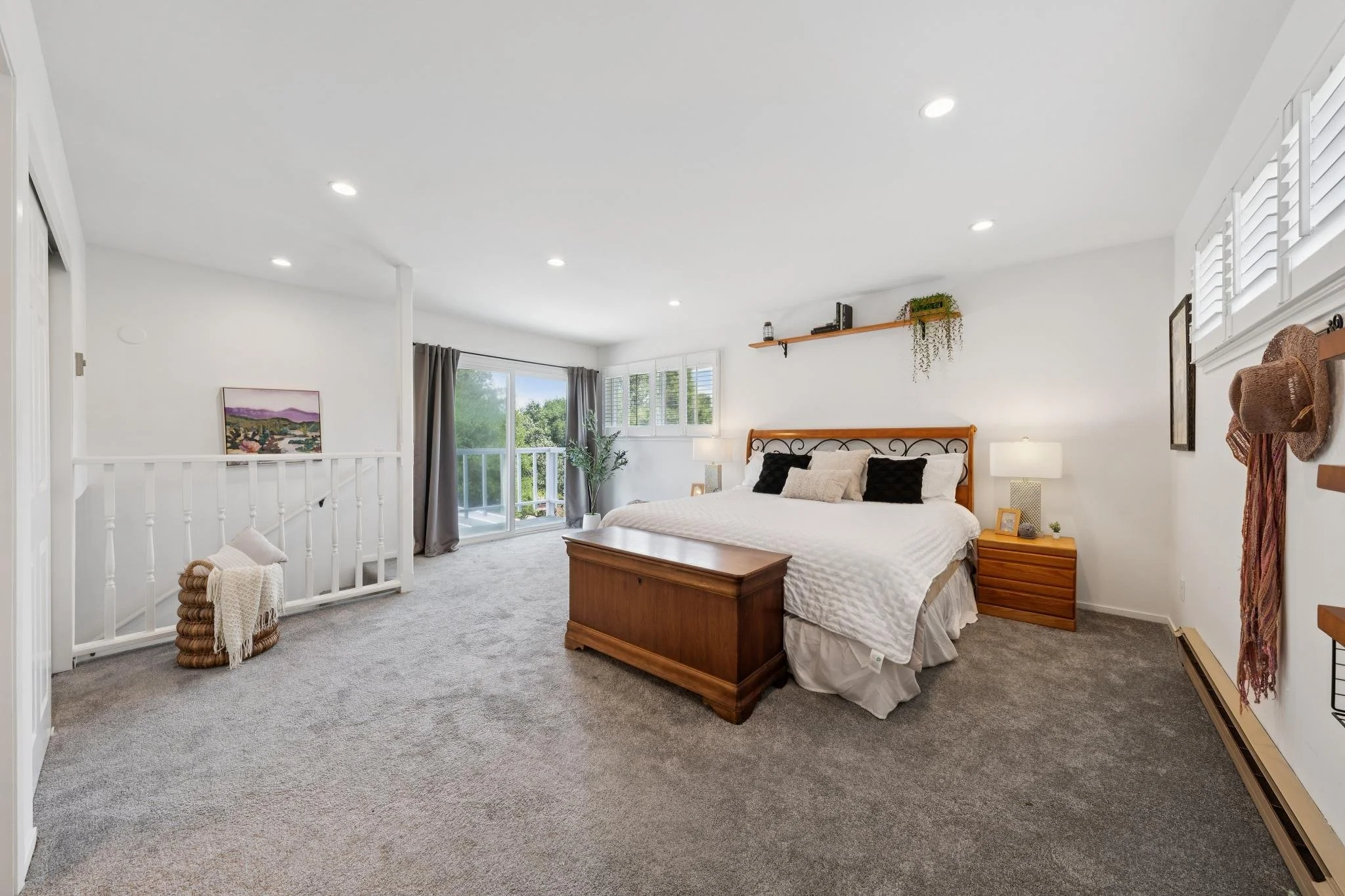 A spacious bedroom with a large bed, nightstands, window with shutters, and a balcony door, decorated with wall art and plants.