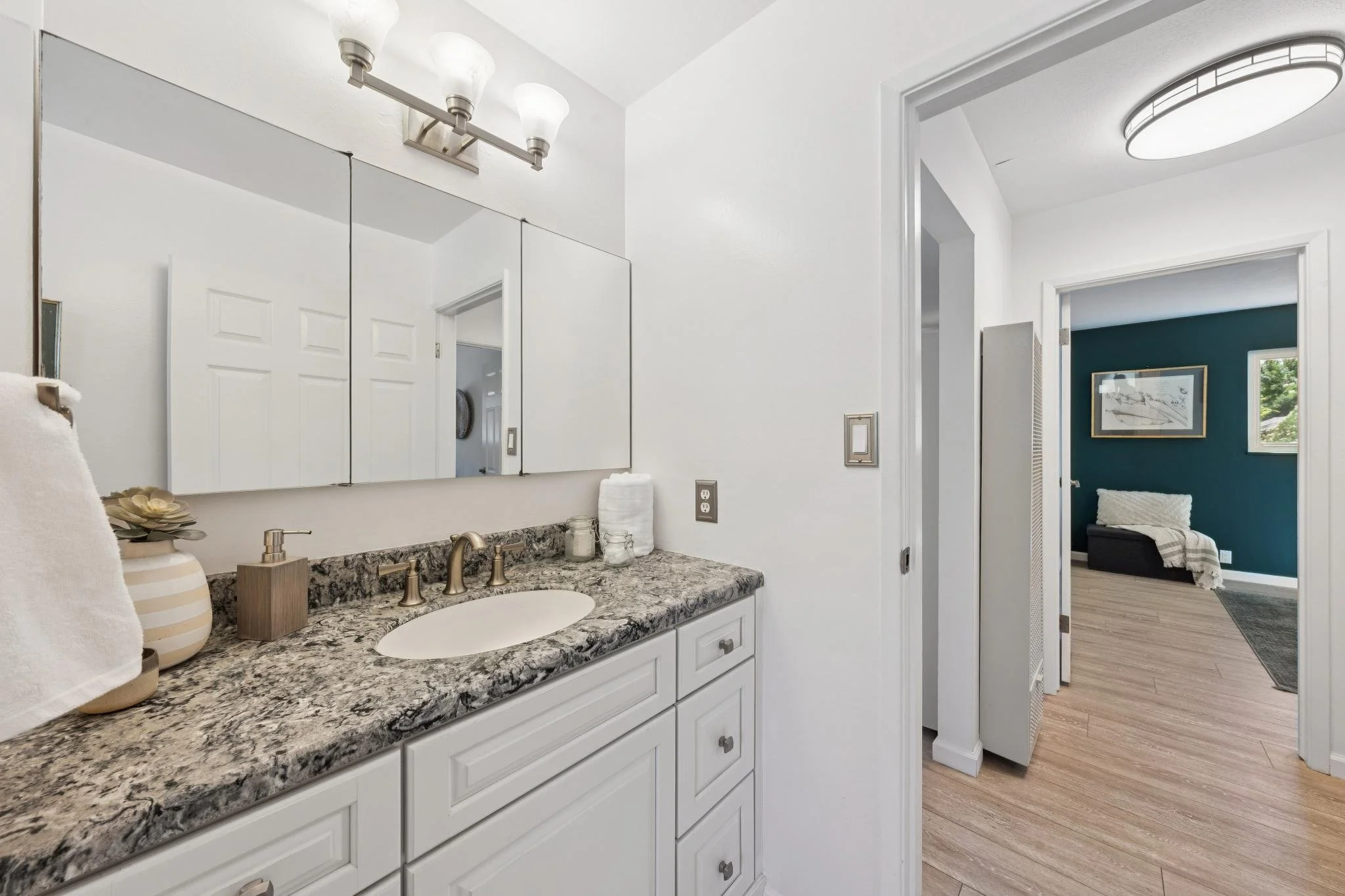 Bathroom vanity with granite countertop, sink, and soap dispenser, mirror above with three light fixtures, towel, and decorative vase, view into adjacent room with blue wall and artwork.