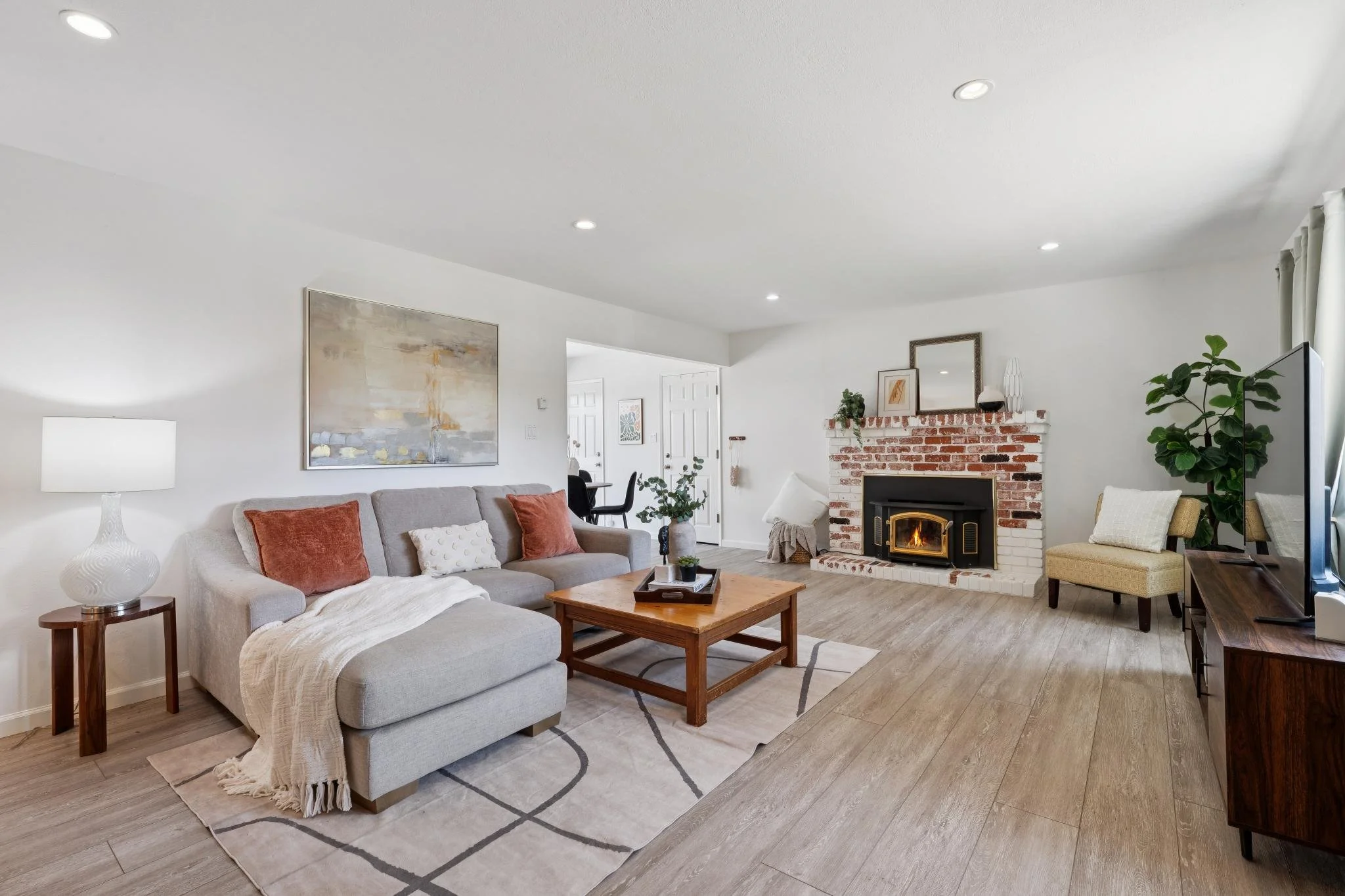 Living room with a beige sofa, wooden coffee table, fireplace, and flat-screen TV, with plants and artwork on a white wall.