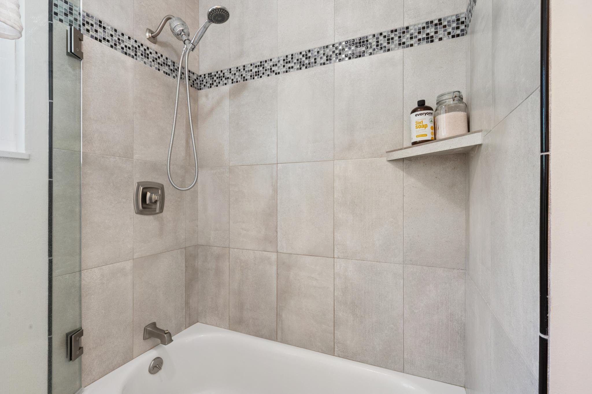 A shower area with beige tiled walls, a decorative black and gray mosaic tile border, and a built-in shelf holding soap and bath salts. The shower has a handheld showerhead and a wall-mounted control.