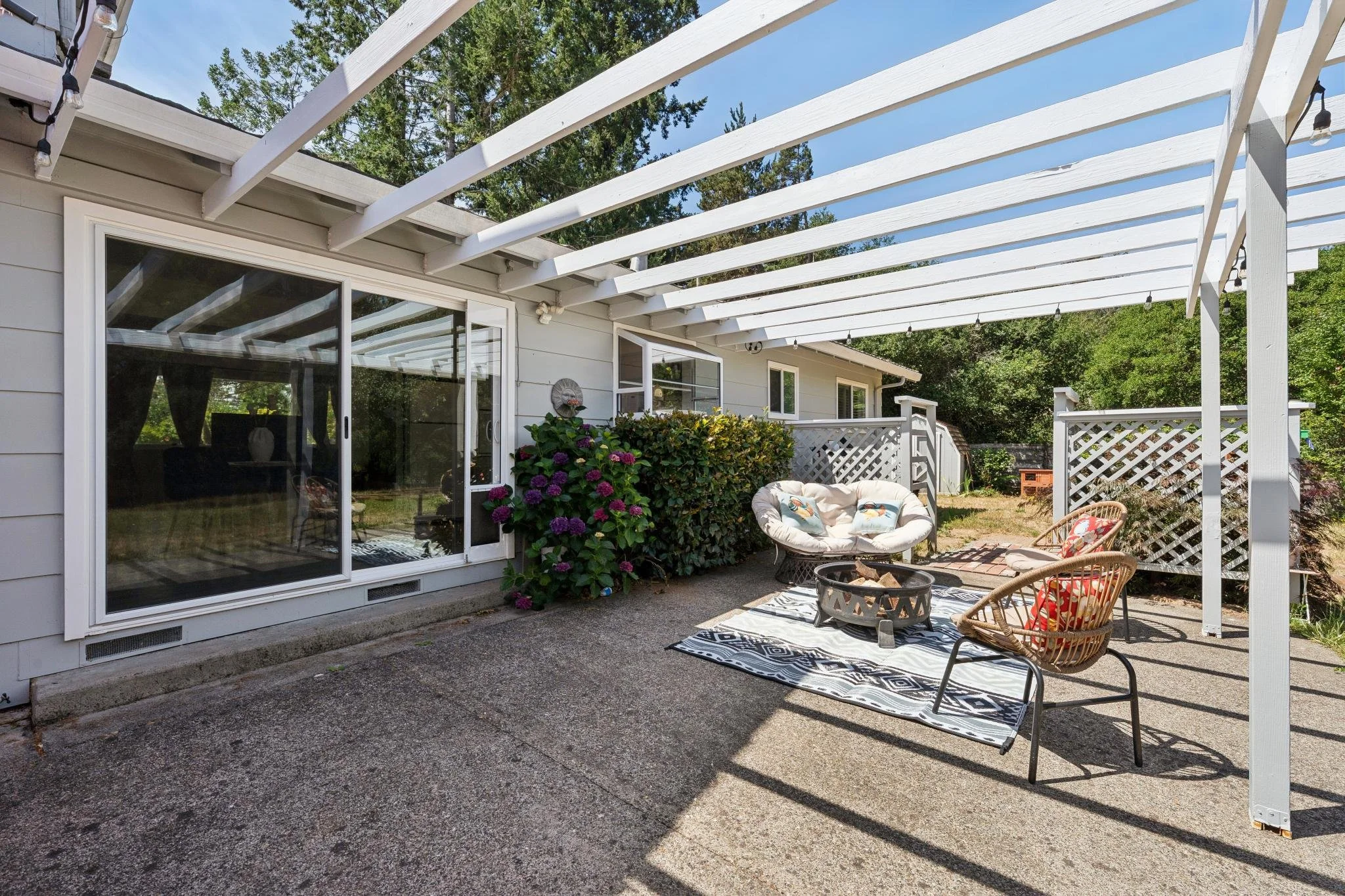 Backyard patio with white pergola, sliding glass door, bushes, outdoor seating including chairs and small table, and string lights