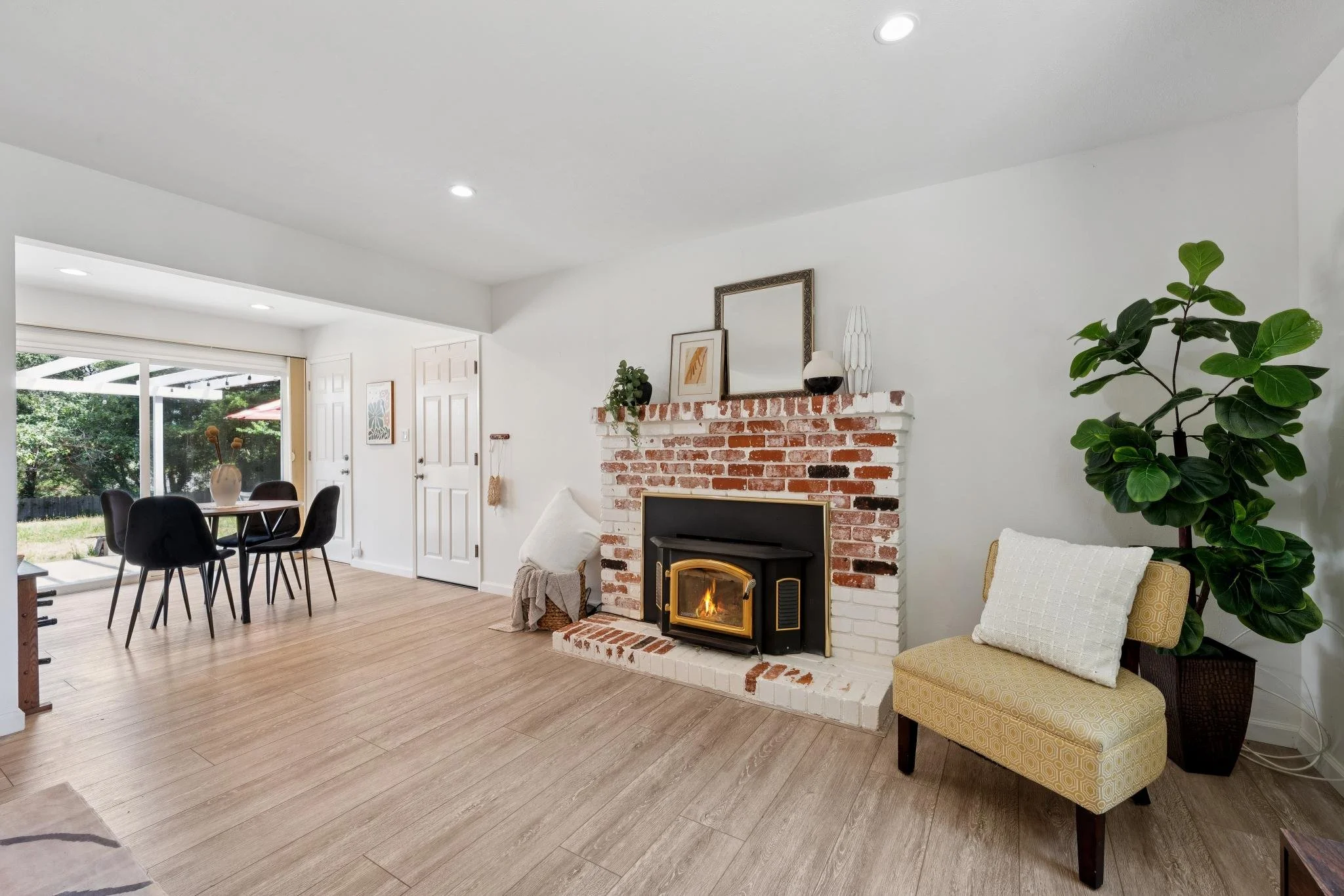 Living room with a brick fireplace, a yellow patterned armchair, a large green houseplant, dining table with black chairs, and sliding glass door opening to outdoor patio.