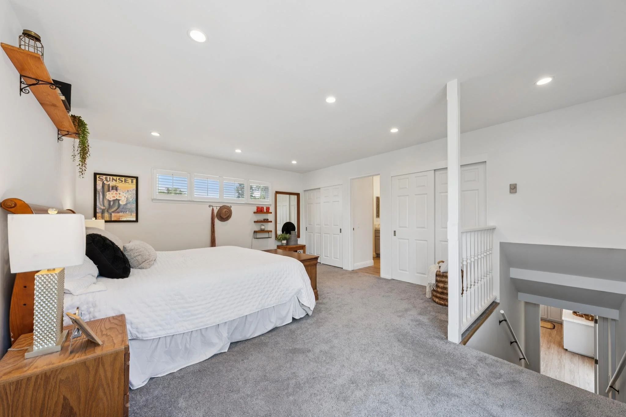 A bright, modern bedroom with a large bed with white bedding, a wooden nightstand, a wall art piece labeled "Sunset," and a window with white shutters. The room has white walls, a ceiling with recessed lighting, and a carpeted floor. A staircase with
