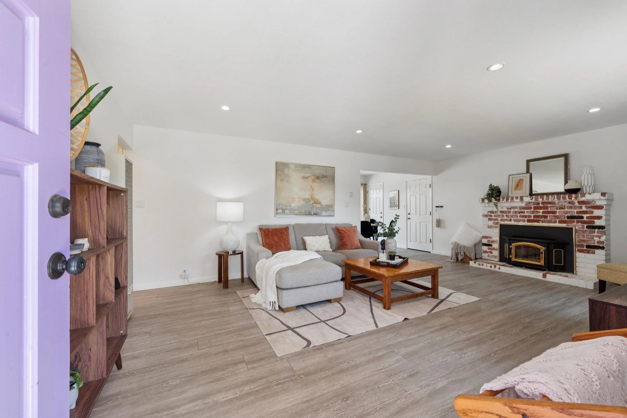 Living room with a gray sofa, wooden coffee table, white and brick fireplace, framed artwork, table lamp, and decorative items.