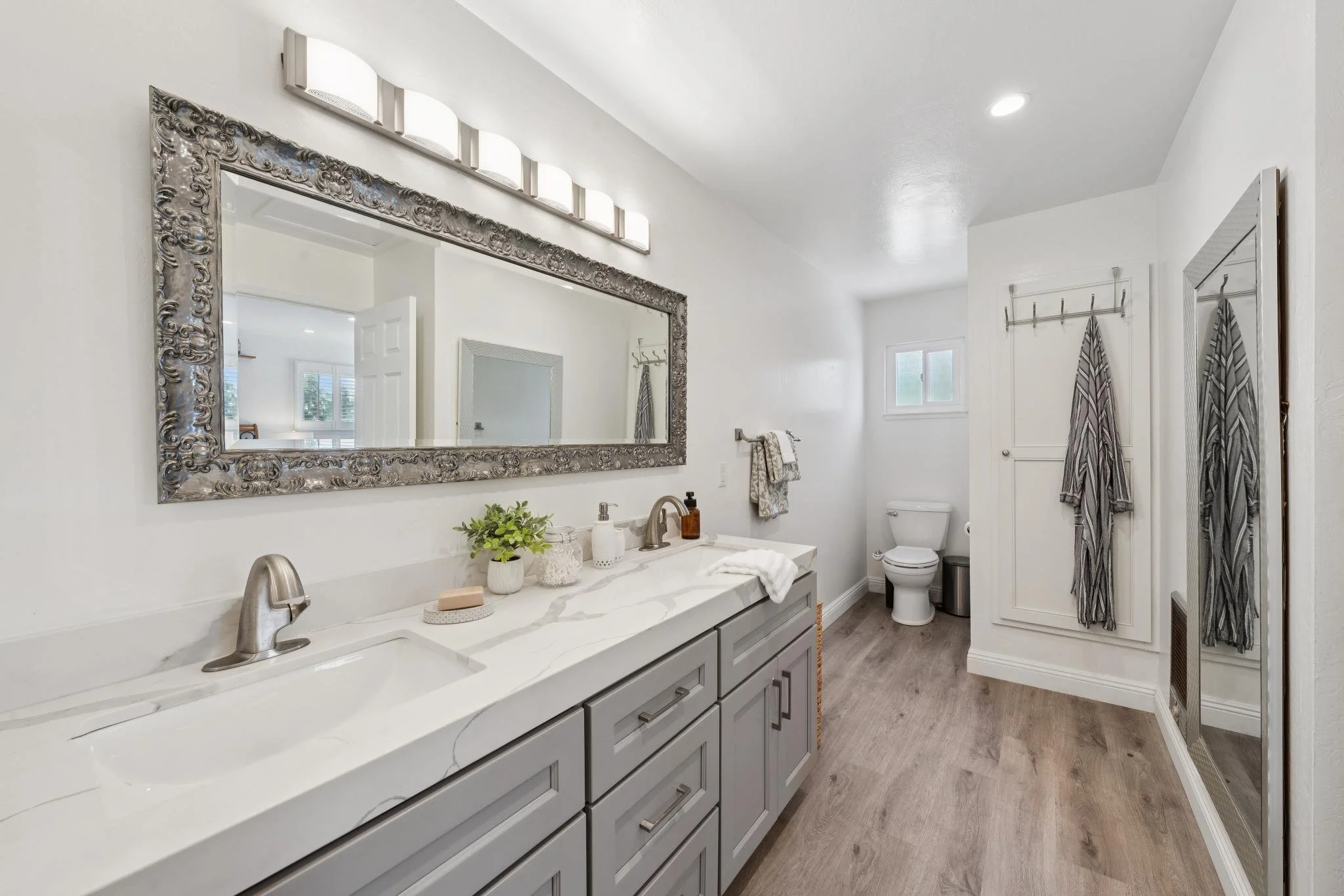 A modern bathroom with a white vanity, marble countertop, and large decorative mirror. There are two sinks with silver faucets, a plant, soap dispenser, and towel on the counter. The bathroom has white walls, wooden flooring, and a window with natura