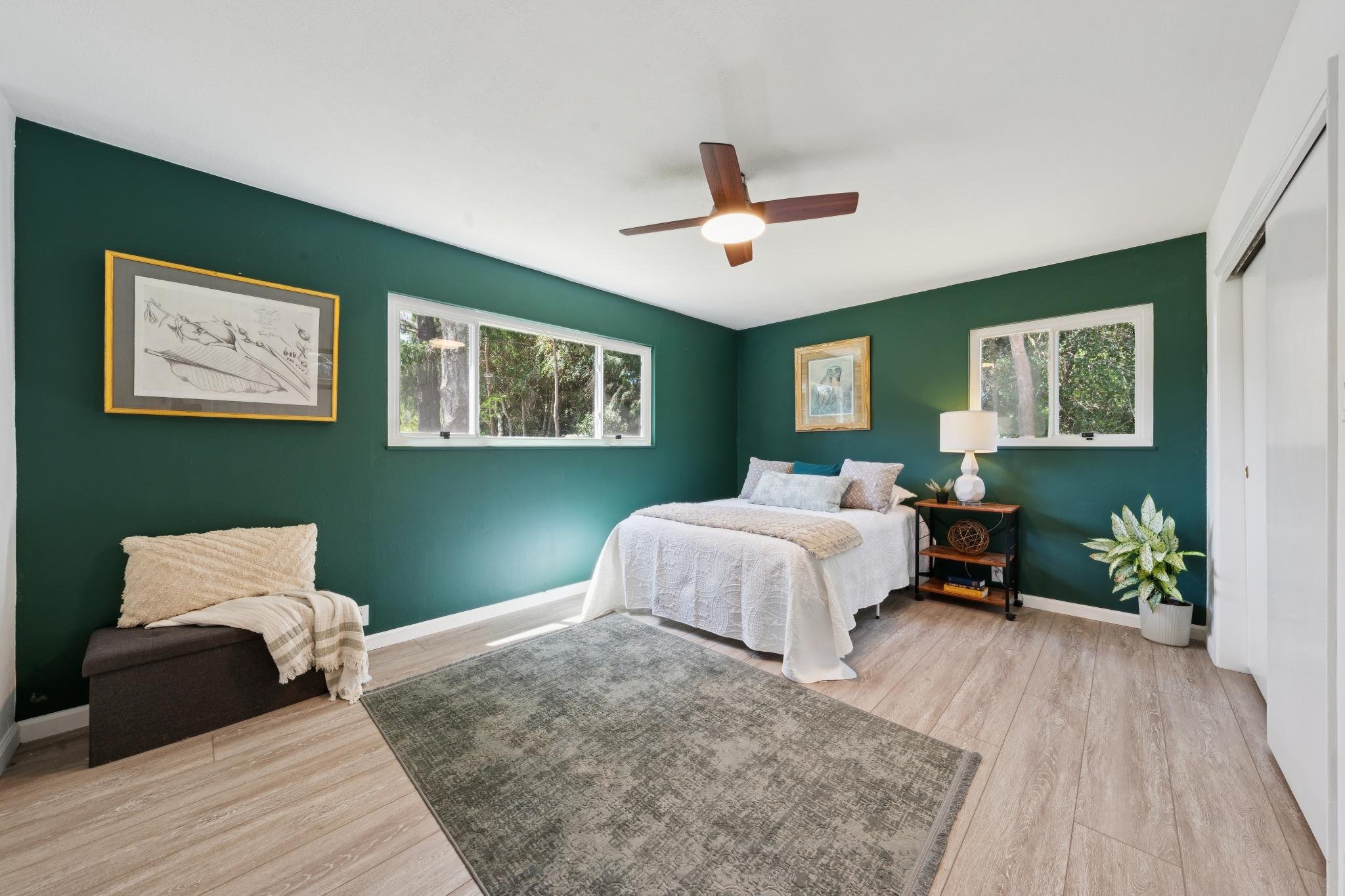A bedroom with green walls, two windows, a bed with white bedding and pillows, a nightstand with a white lamp, a framed picture on the wall, a potted plant, and a ceiling fan.