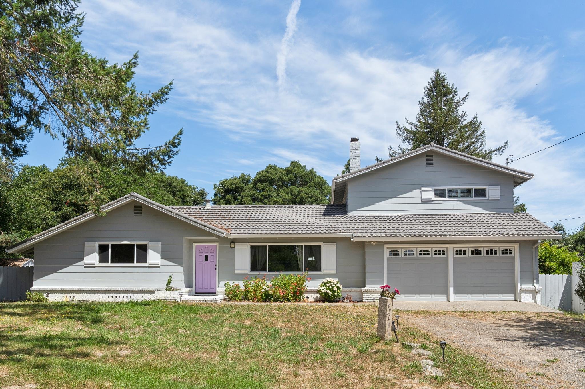 A house with a gray exterior, pink front door, and a white garage door, surrounded by a lawn with flowers. Trees and a blue sky with light clouds are in the background.