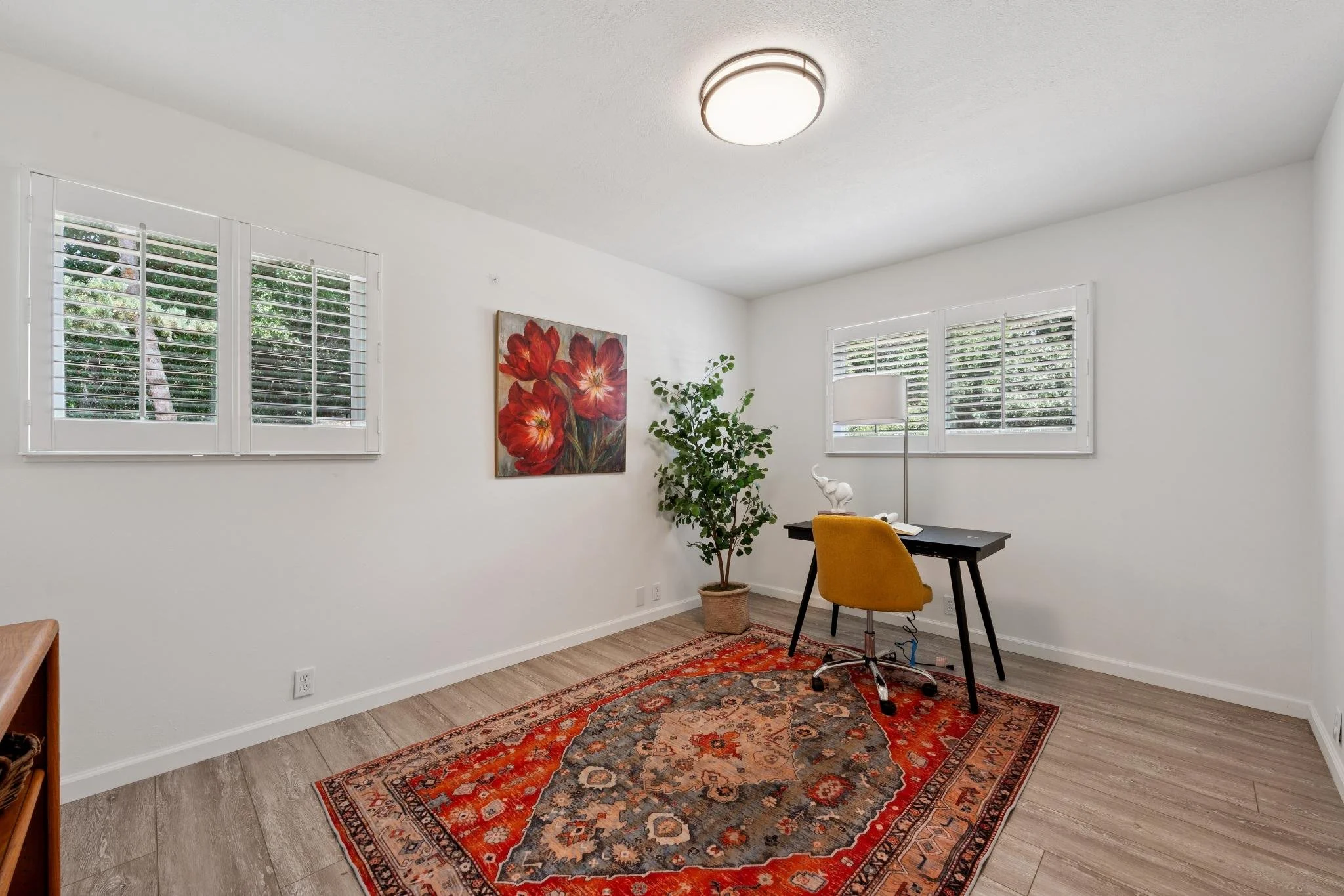 A bright, minimalist home office with white walls, two windows with white shutters, a colorful rug, a potted plant, a wall art of red flowers, a yellow chair, a black desk, and a floor lamp.