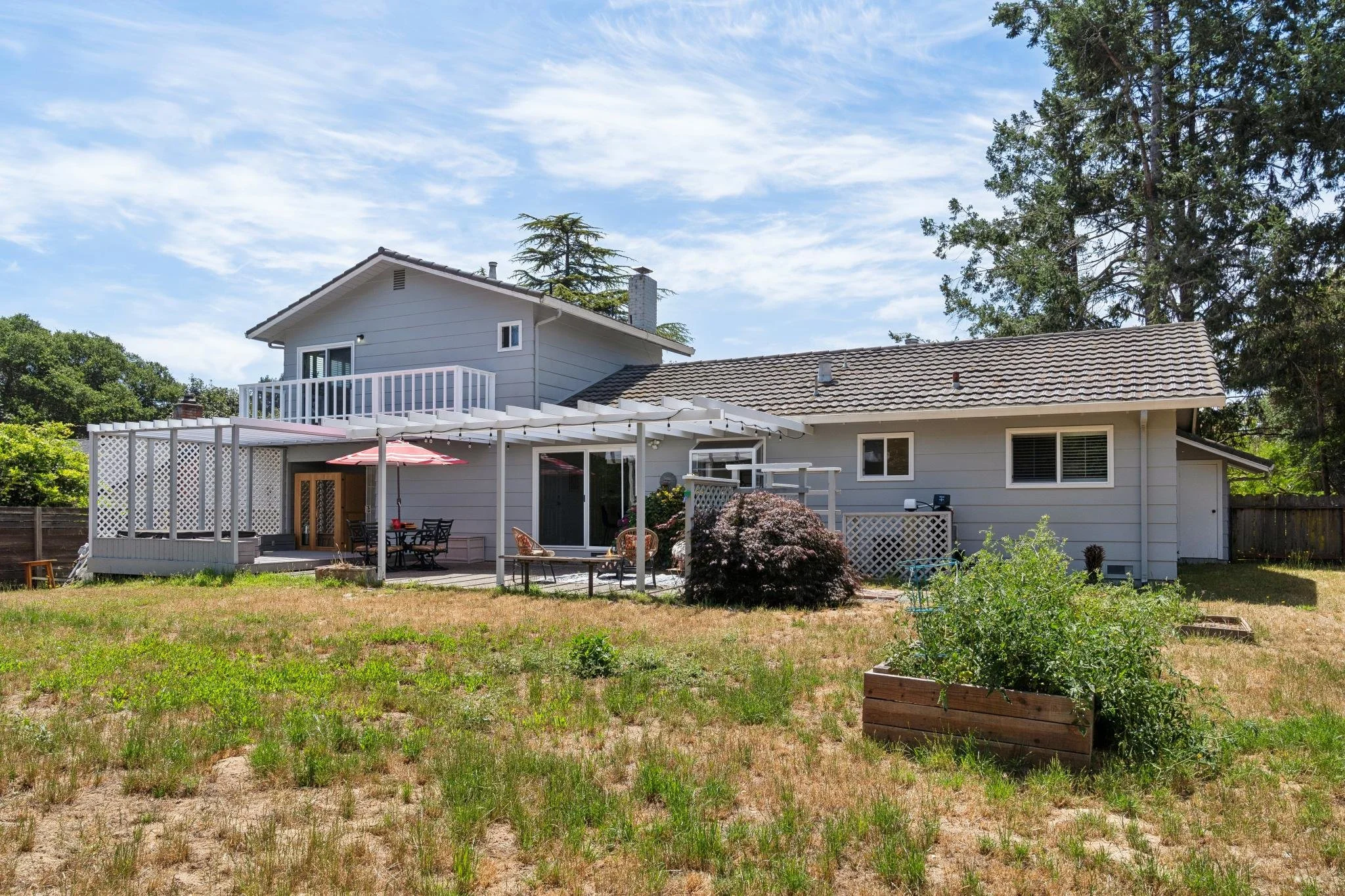 Backyard view of a two-story house with a patio, garden, and trees in the background on a sunny day.