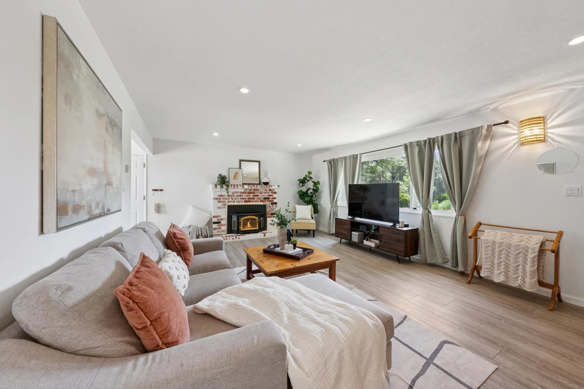 Living room with a beige sofa with throw pillows, a wooden coffee table, a fireplace with a brick surround, a TV on a wooden stand, large windows with curtains, and a wall-mounted light fixture.