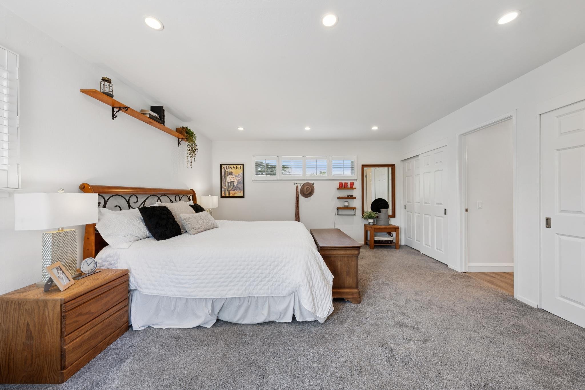 A bedroom with a wooden bed frame, white bedding, and multiple pillows. There are wall decorations including a framed sunset picture, wall shelves with decor, and a mirror. The room has windows with white blinds, a side table with a clock and framed 
