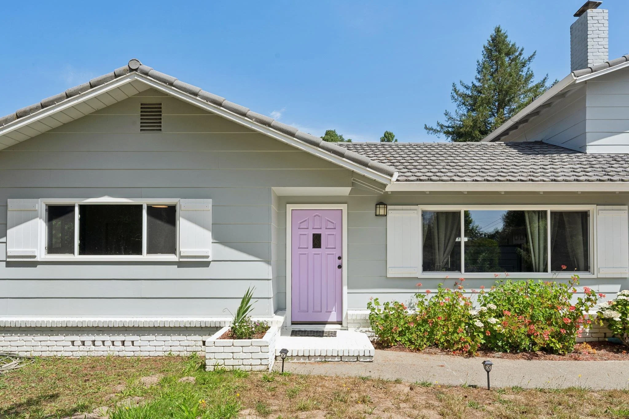 Front of a house with a purple front door, two large windows, white siding, and a front garden with flowers and bushes under a clear blue sky.