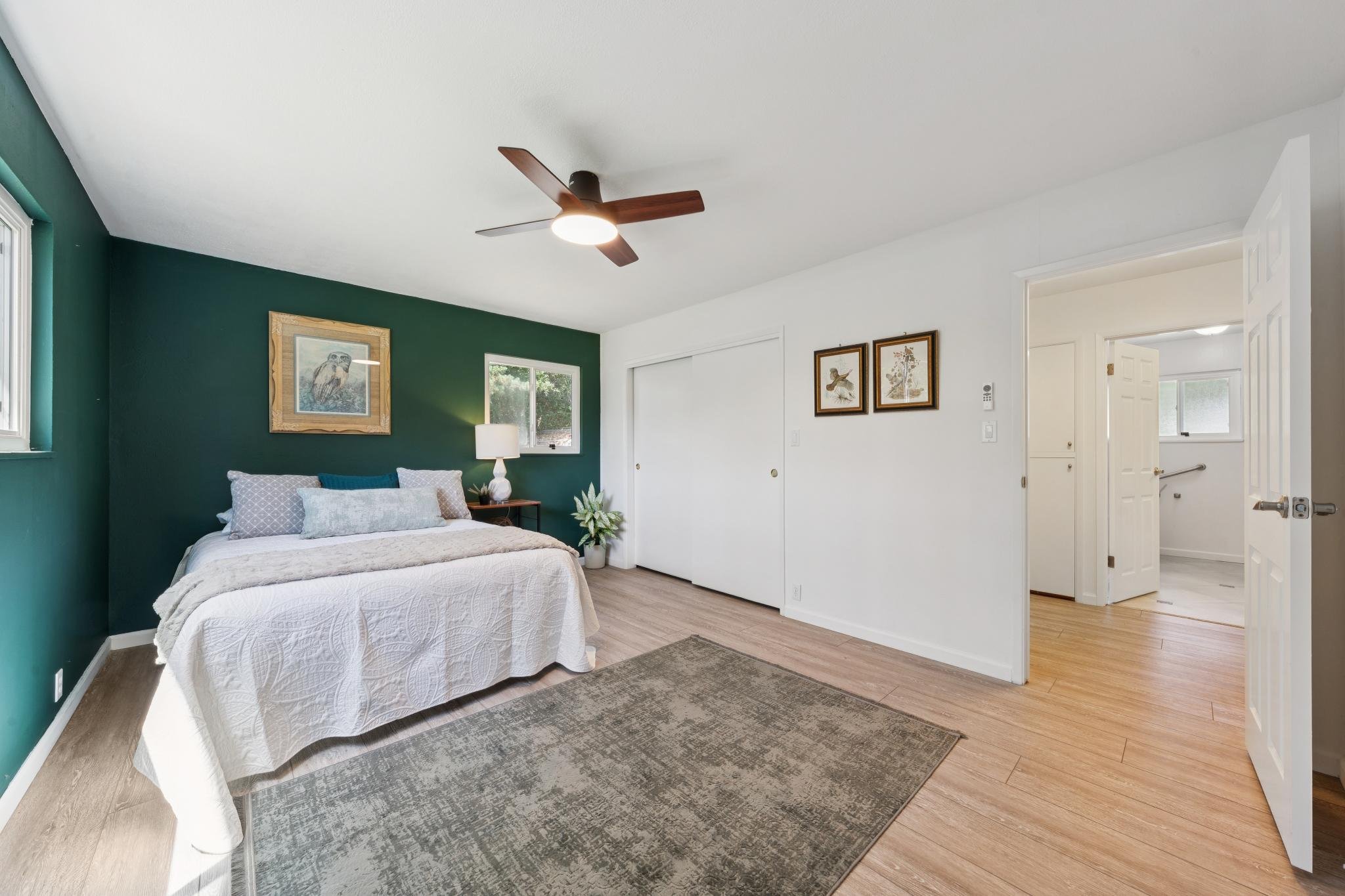 Bedroom with white bed, green accent wall, framed bird and owl pictures, window, white door, ceiling fan, and wooden floors.
