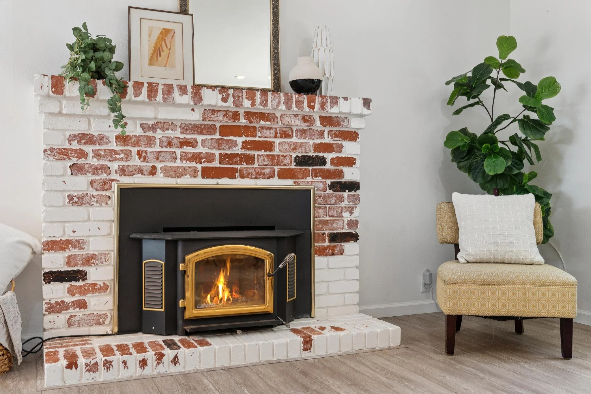 Living room with a white and red brick fireplace, a mirror, framed artwork, a potted plant, and a beige chair with a patterned pillow.