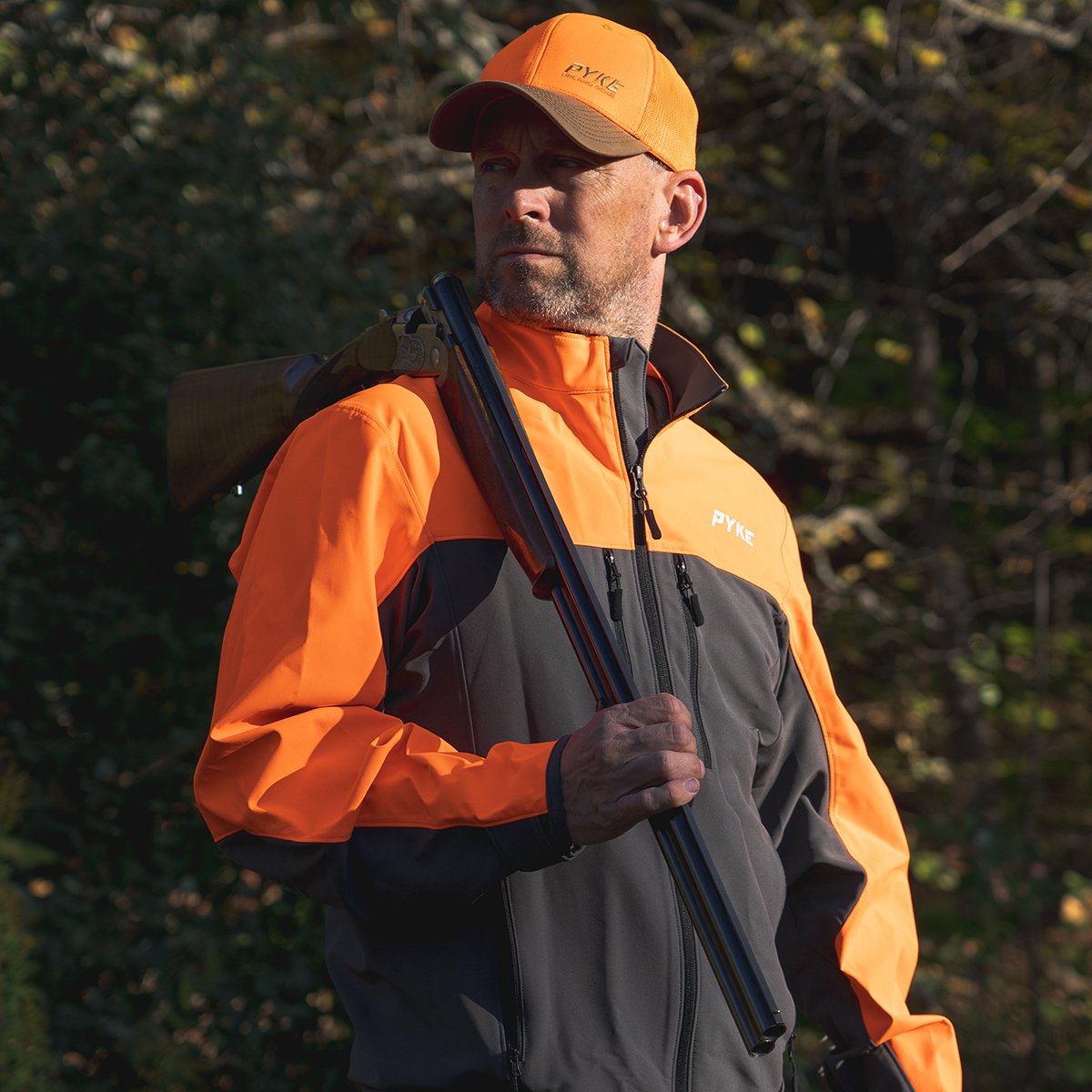Man in orange and black outdoor jacket and cap carrying a shotgun over his shoulder in a wooded area.