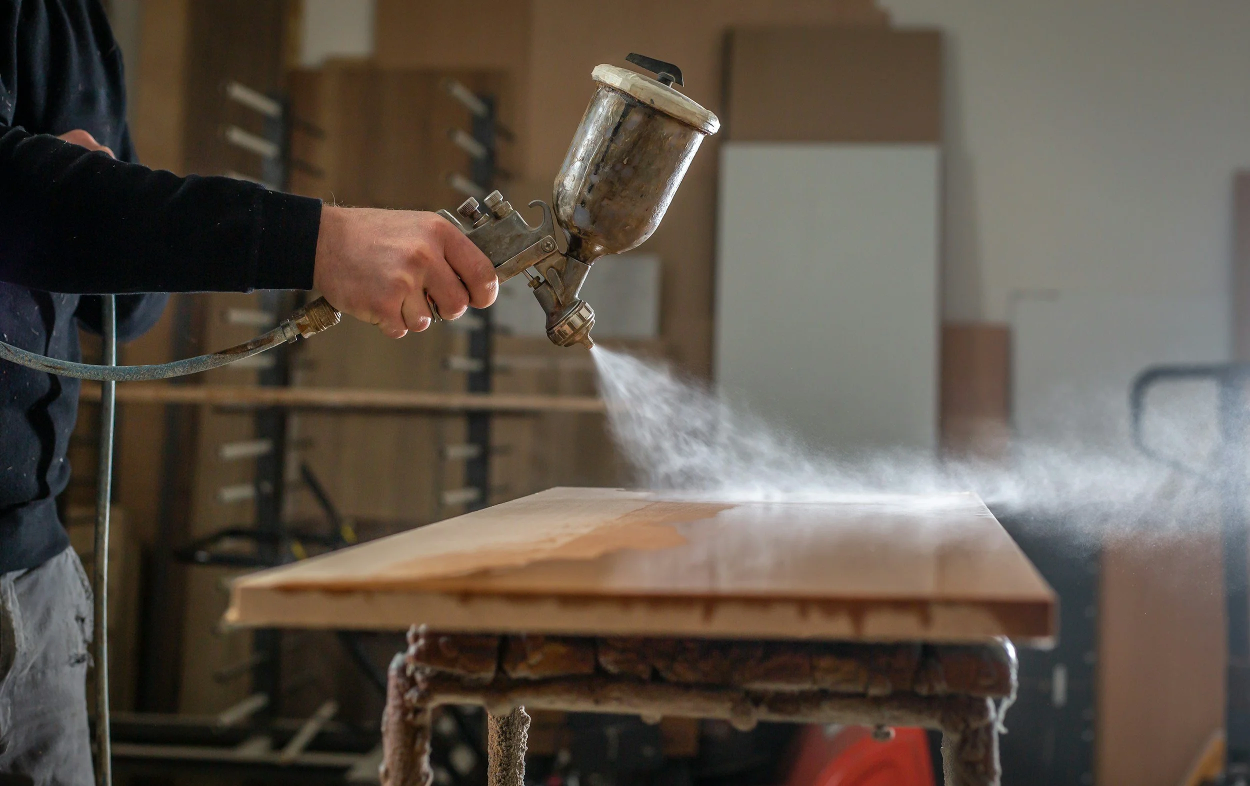 Person holding a spray gun and applying a finish to a wooden surface in a workshop.